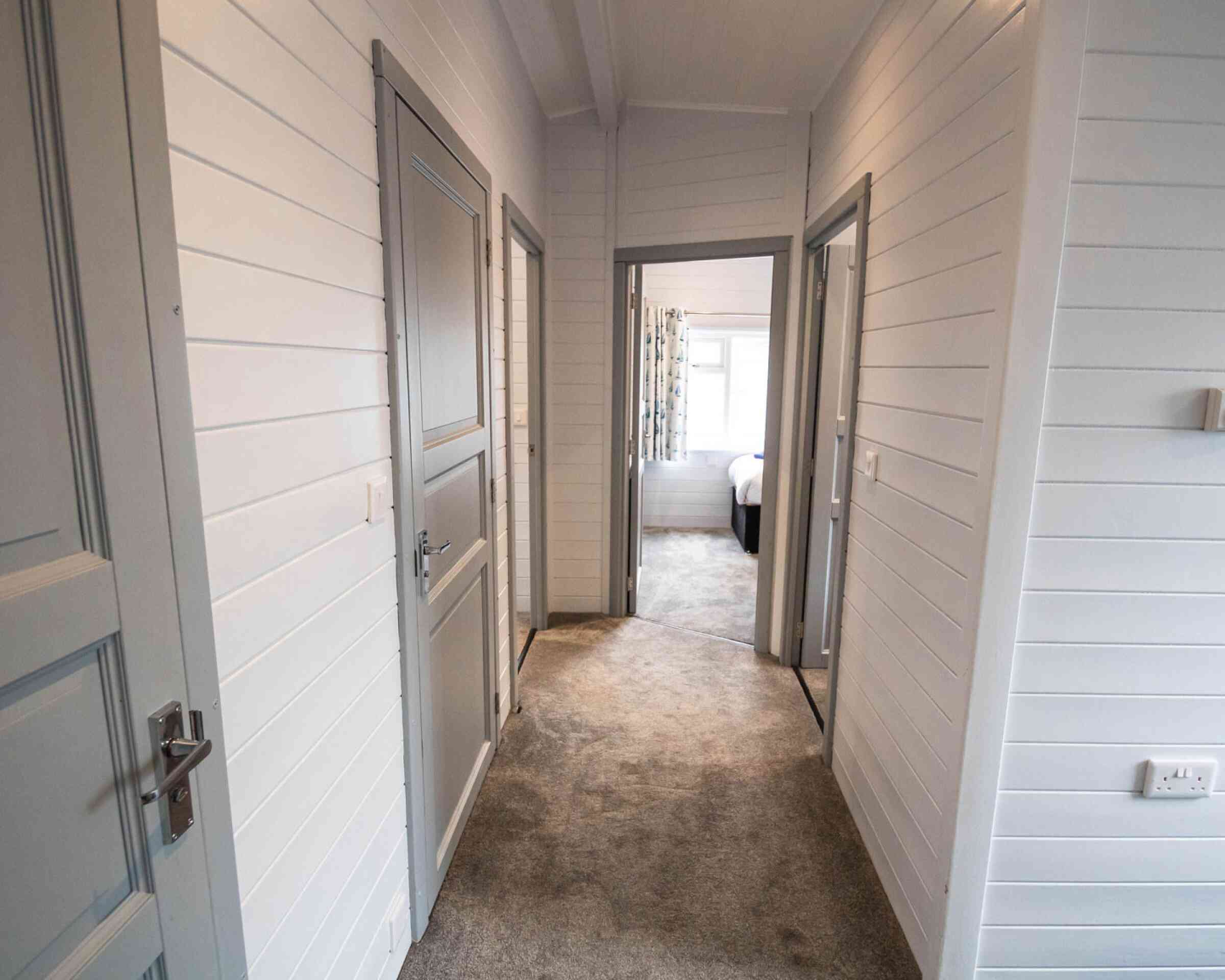 A well-lit hallway featuring light gray paneling and beige carpet. There are three doorways leading to different rooms, with a glimpse of a bedroom at the end. The walls are white with a clean and modern aesthetic.