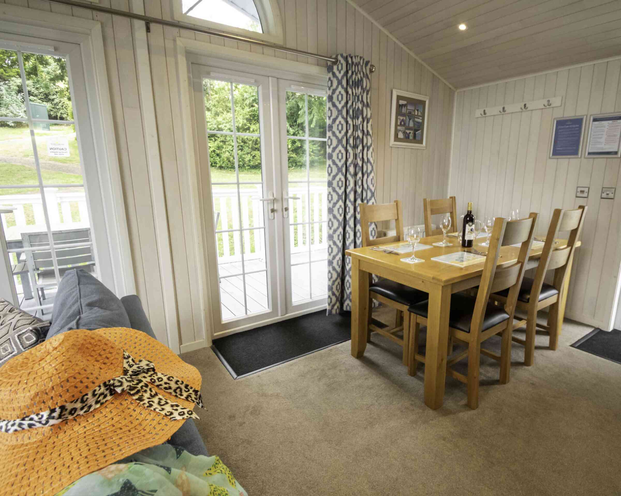 Bright and airy dining area with a wooden table set for six, surrounded by plush chairs. A cozy sofa is on the left with a large straw hat resting on it. French doors lead to an outdoor view, and decorative curtains frame the windows.