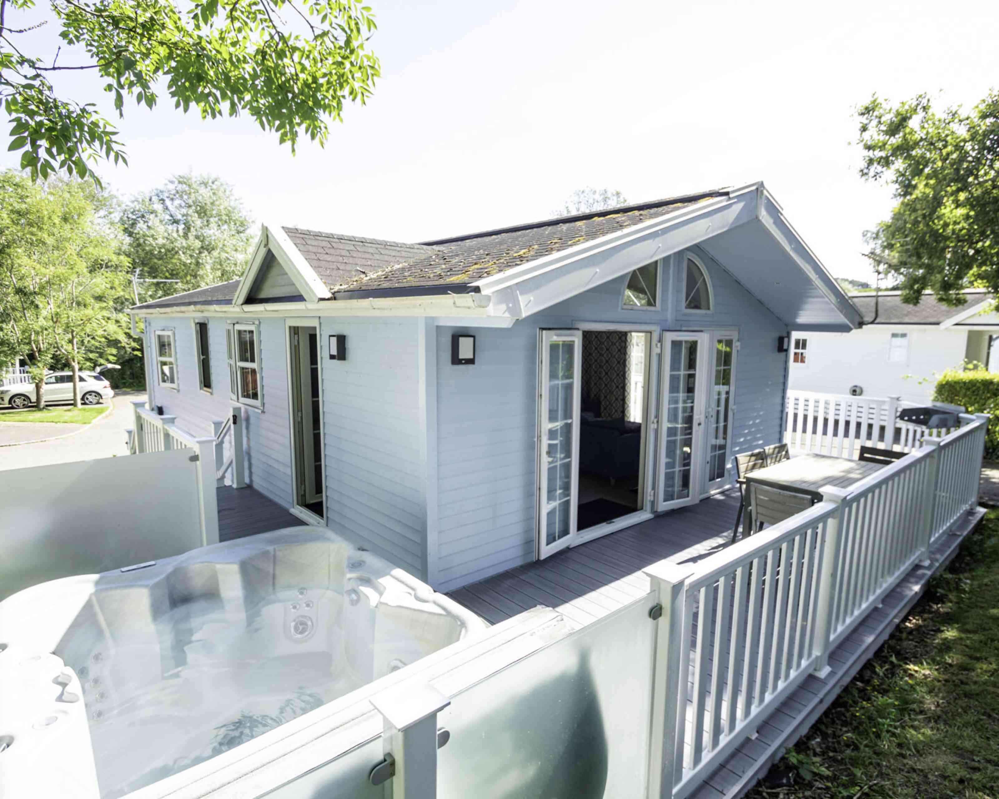 A light blue wooden cabin with large windows and double doors. A hot tub is visible on the deck in front of the cabin. Lush greenery surrounds the area, and there are cars parked nearby.