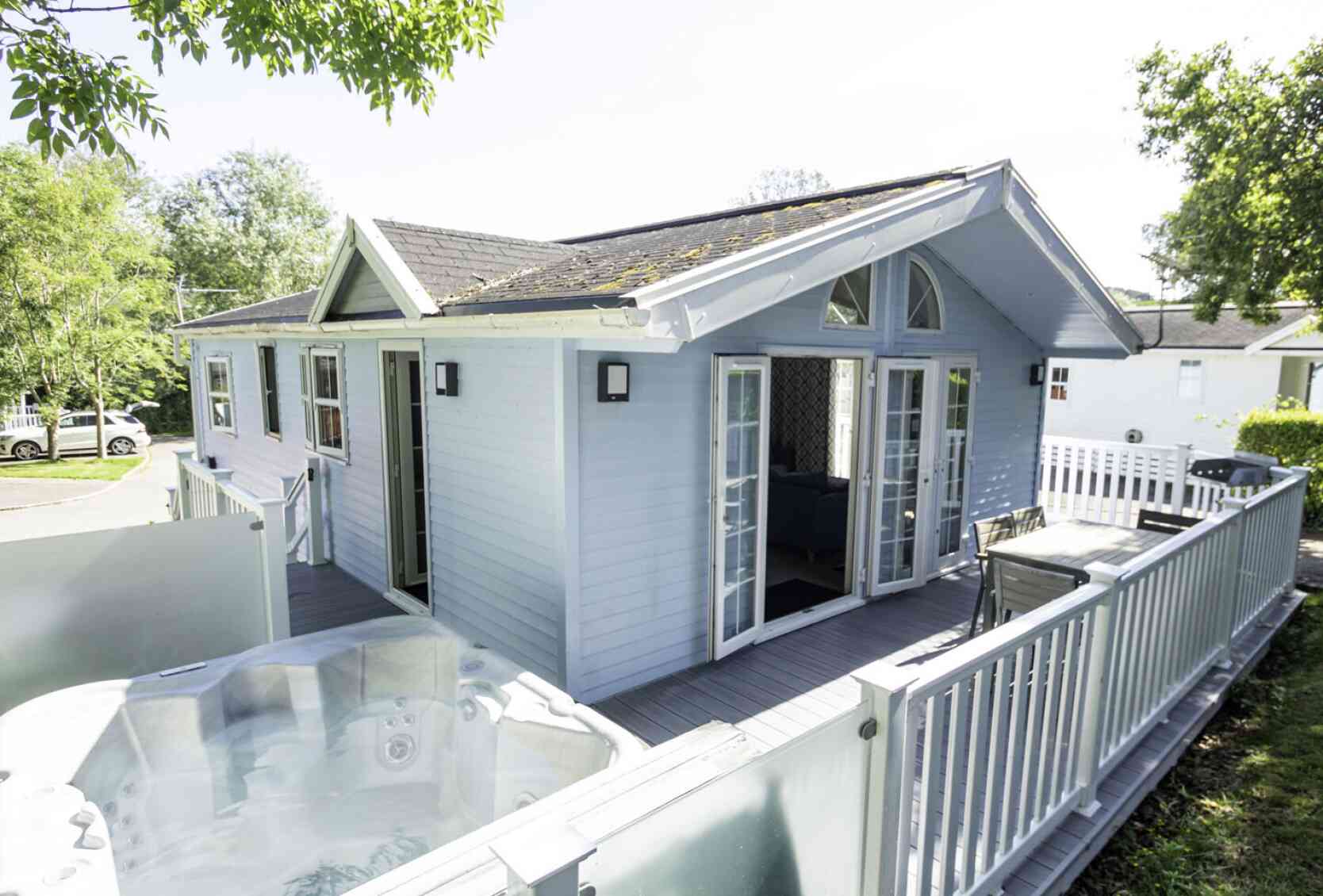 A light blue wooden cabin with large windows and double doors. A hot tub is visible on the deck in front of the cabin. Lush greenery surrounds the area, and there are cars parked nearby.