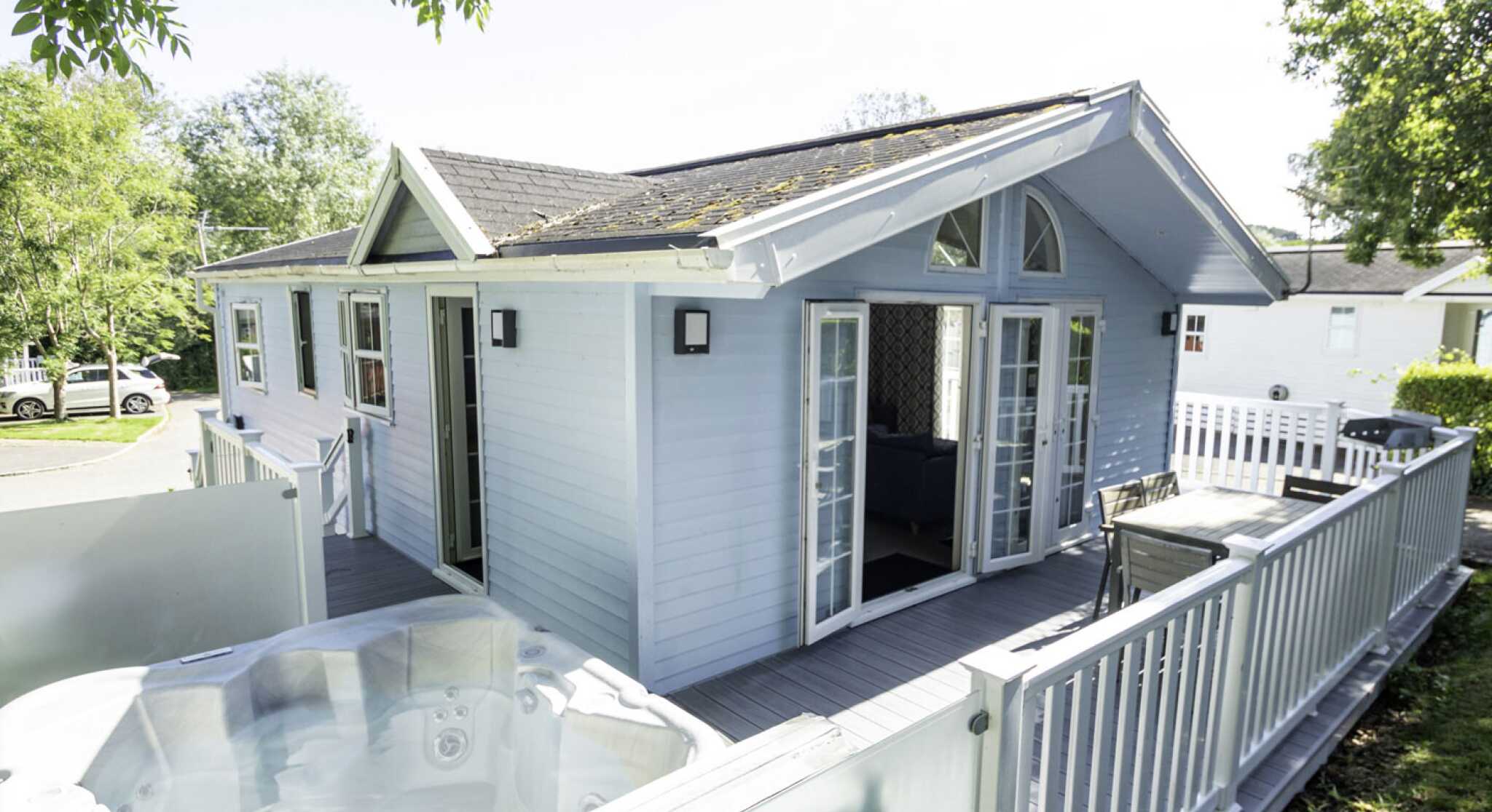 A light blue wooden cabin with large windows and double doors. A hot tub is visible on the deck in front of the cabin. Lush greenery surrounds the area, and there are cars parked nearby.
