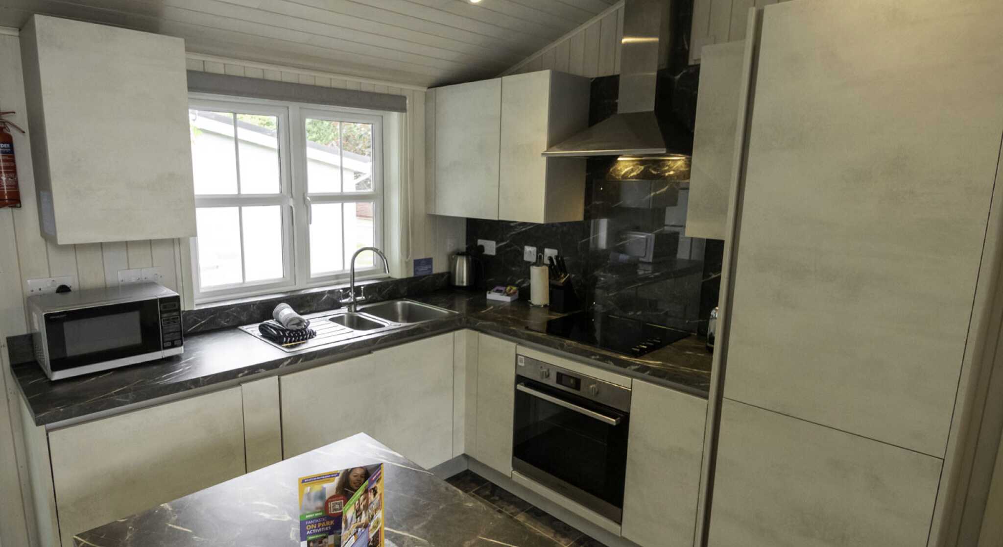 Modern kitchen with sleek white cabinets, a dark marble countertop, and stainless steel appliances. A window provides natural light, and a microwave is on the counter. A bowl and a brochure are placed on the table in the center.