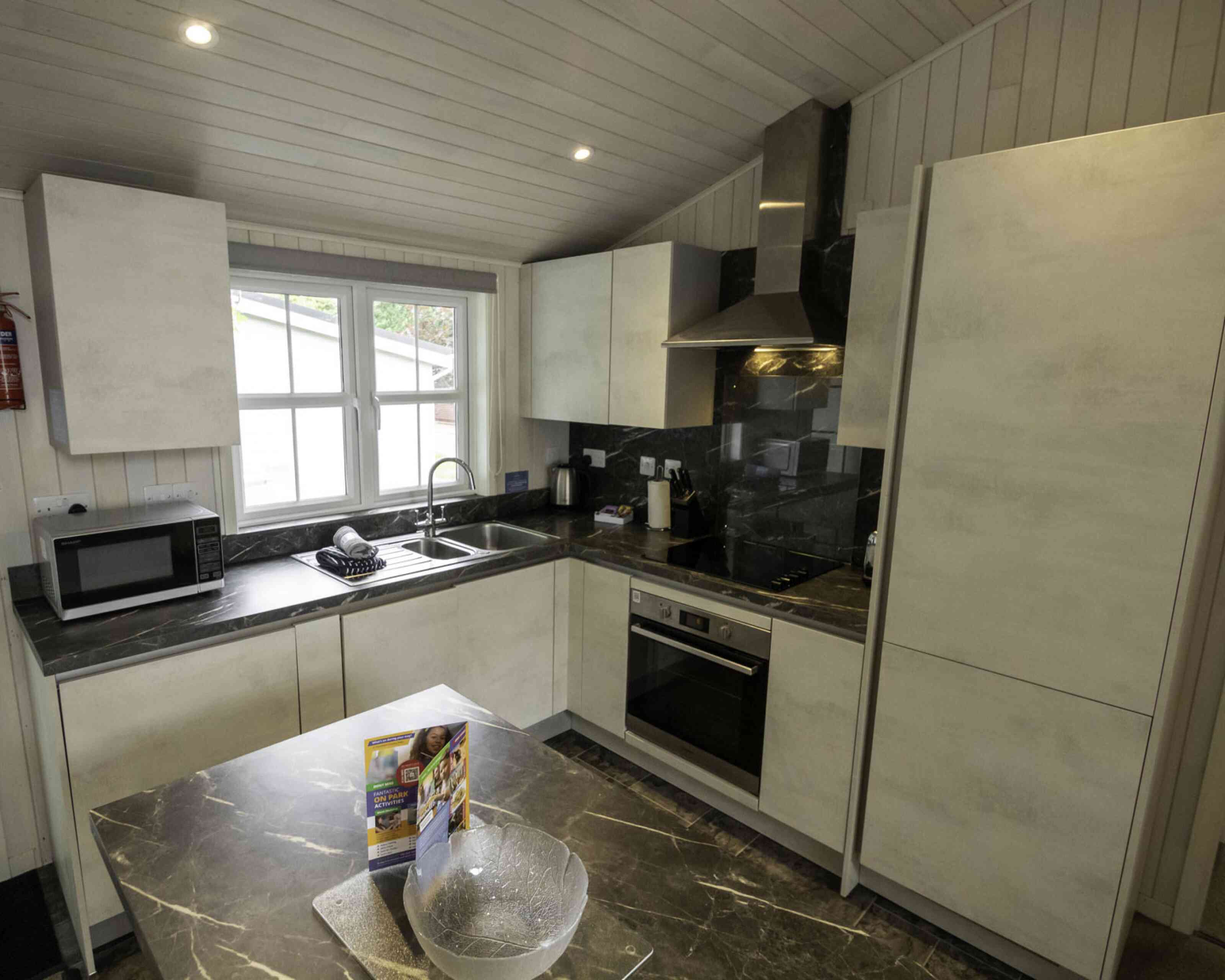 Modern kitchen with sleek white cabinets, a dark marble countertop, and stainless steel appliances. A window provides natural light, and a microwave is on the counter. A bowl and a brochure are placed on the table in the center.