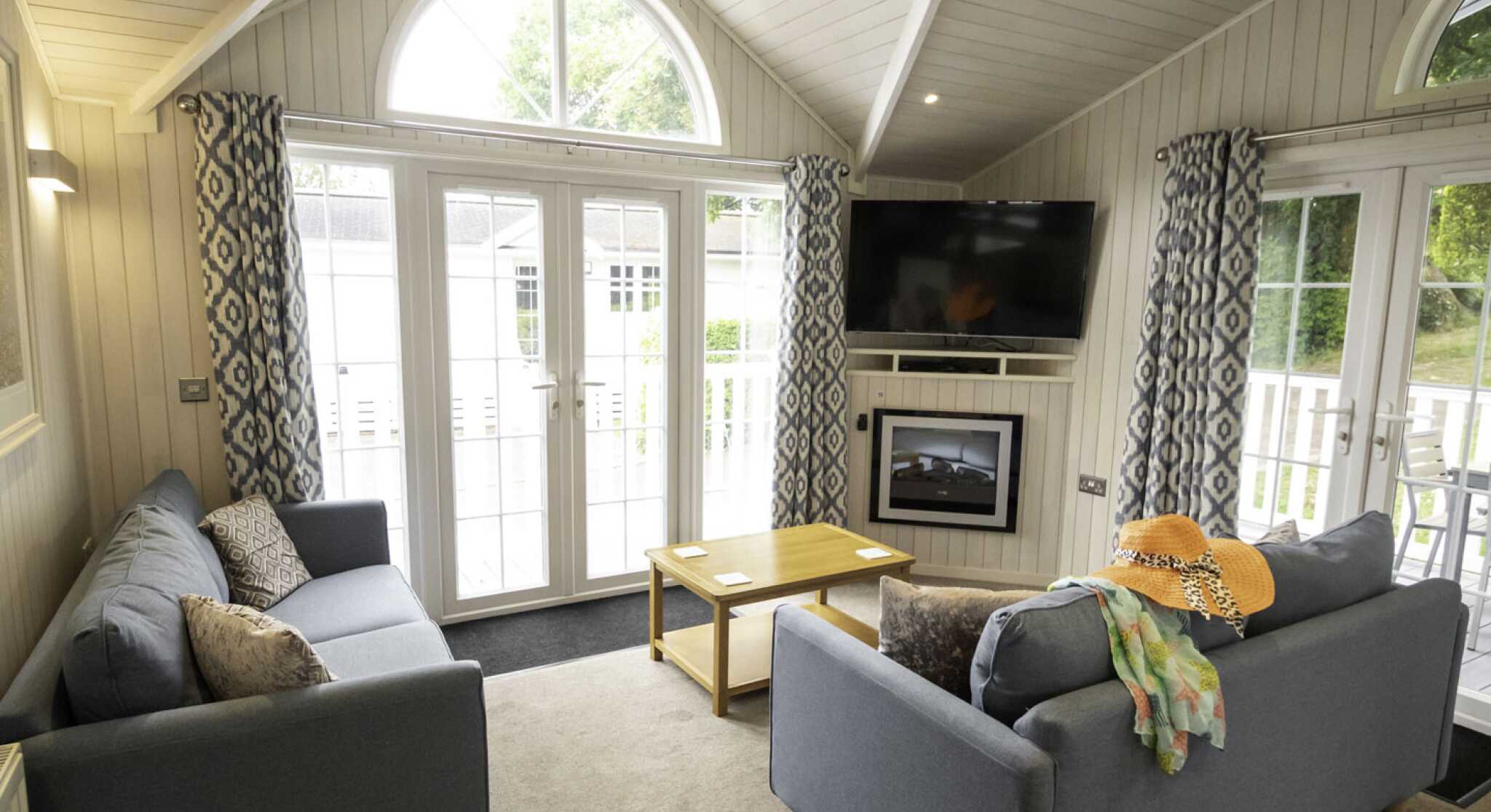 A cozy living room featuring two gray sofas with decorative pillows, a small wooden coffee table, and a flat-screen TV mounted on the wall. Large windows let in natural light, and stylish curtains frame the view. A colorful scarf and a straw hat are casually draped over one sofa.