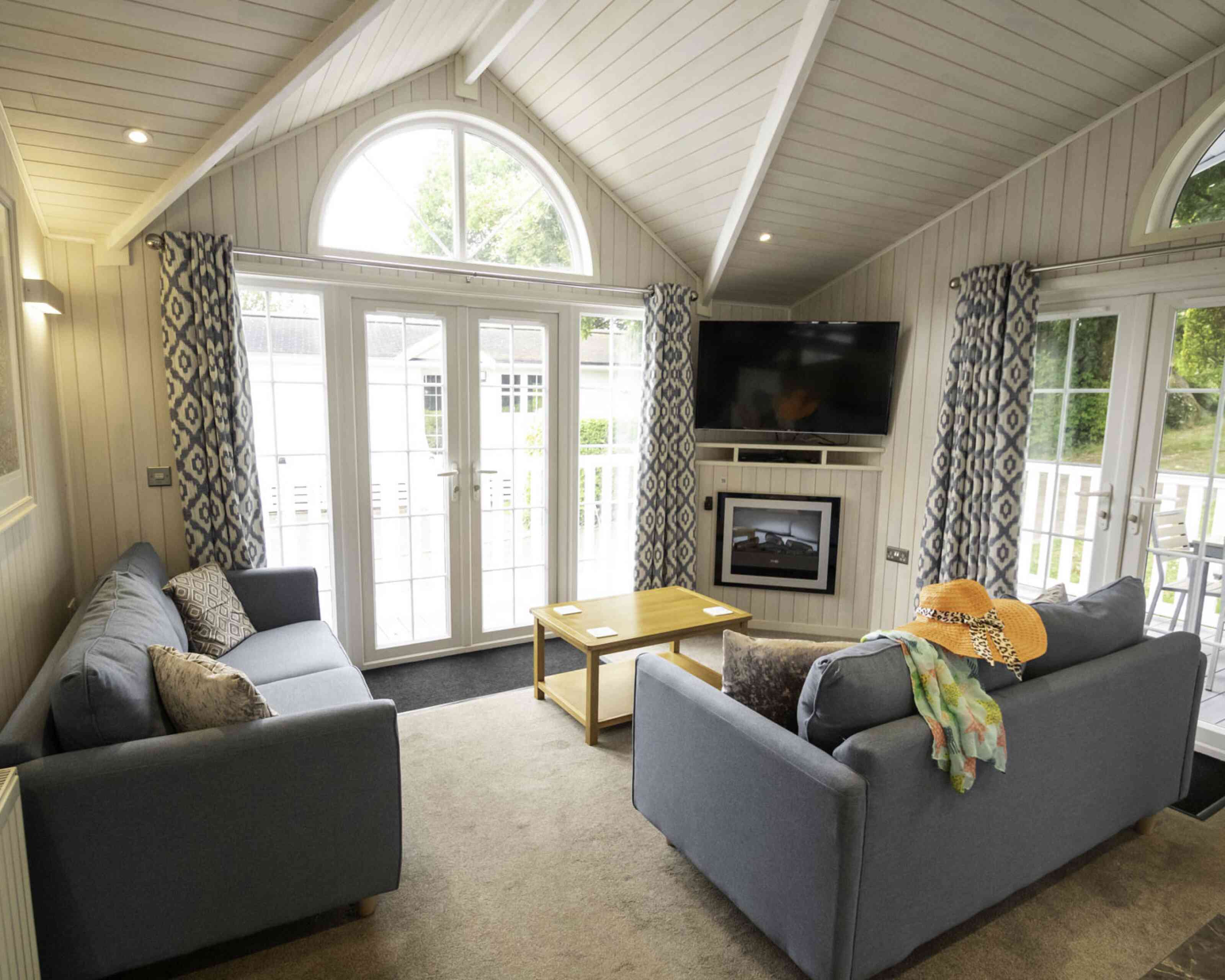 A cozy living room featuring two gray sofas with decorative pillows, a small wooden coffee table, and a flat-screen TV mounted on the wall. Large windows let in natural light, and stylish curtains frame the view. A colorful scarf and a straw hat are casually draped over one sofa.