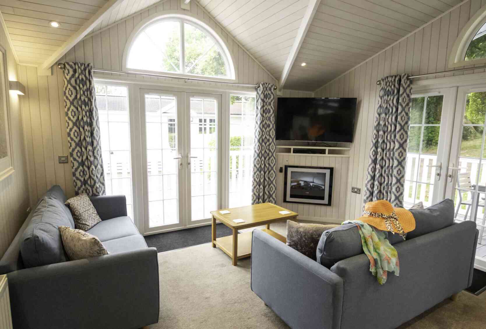 A cozy living room featuring two gray sofas with decorative pillows, a small wooden coffee table, and a flat-screen TV mounted on the wall. Large windows let in natural light, and stylish curtains frame the view. A colorful scarf and a straw hat are casually draped over one sofa.