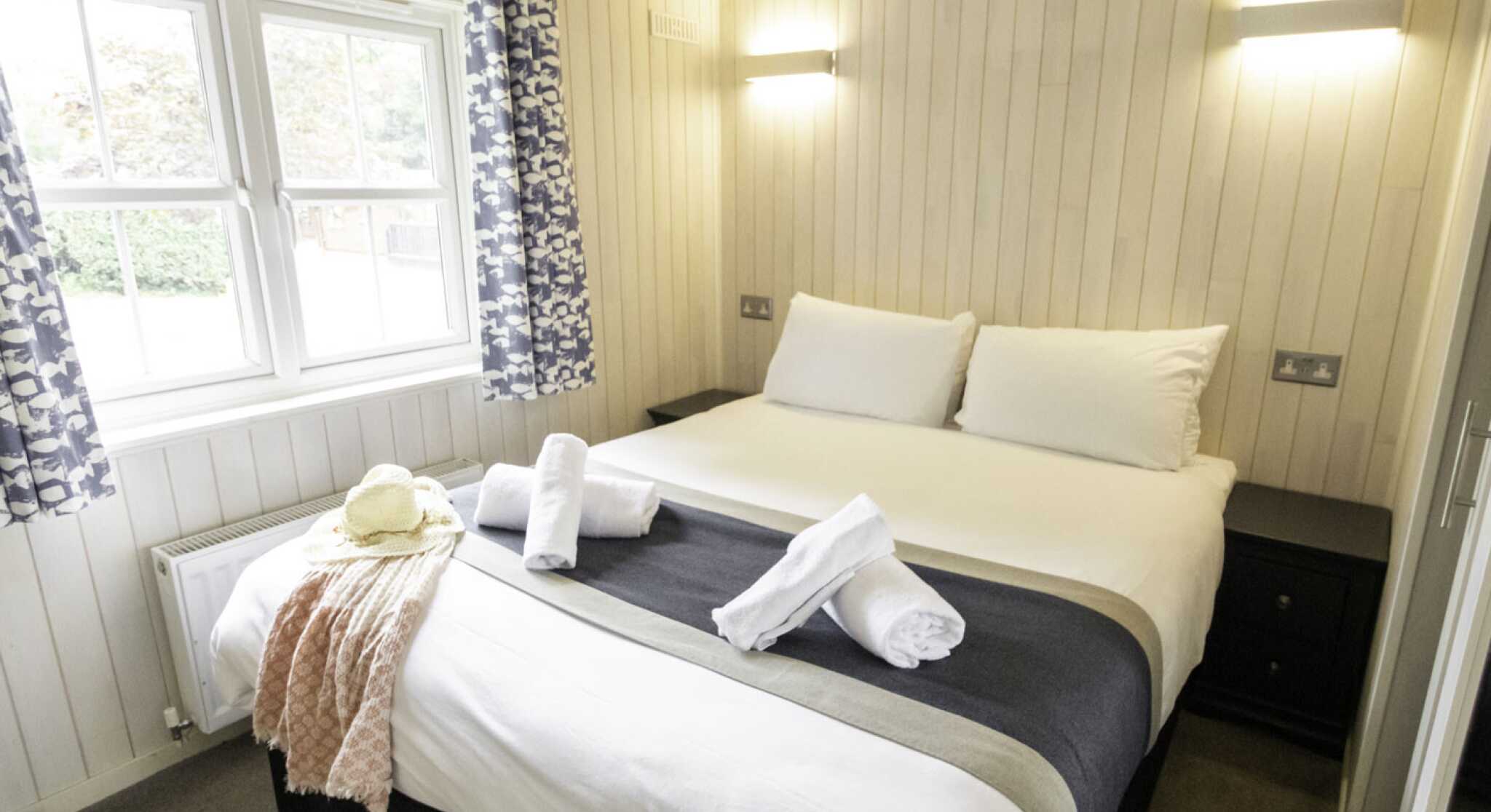 A cozy bedroom featuring a large bed with white linens, gray accents, and neatly arranged towels. Floral curtains frame the window, and soft lighting enhances the warm ambiance. A light peach throw is draped across the foot of the bed.