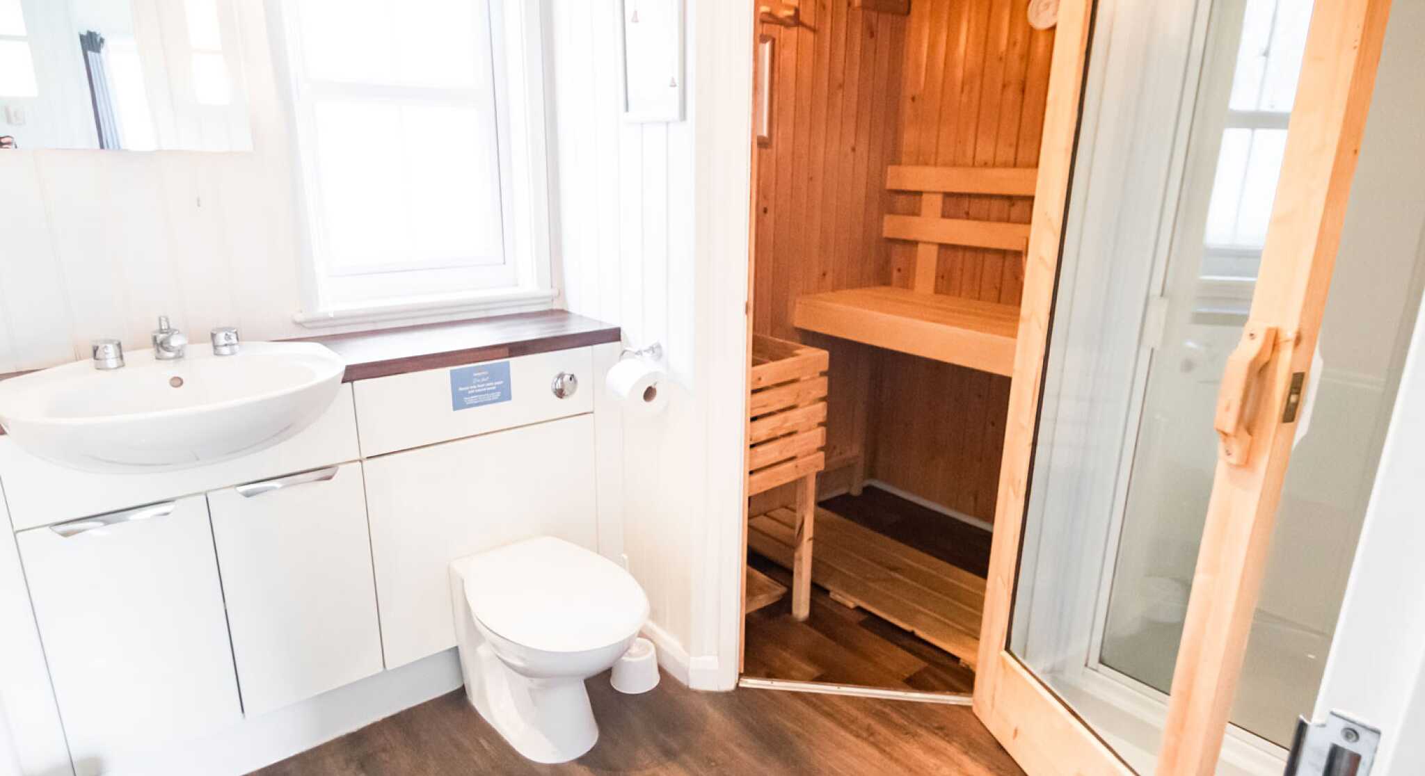 A modern bathroom featuring a white sink, toilet, and a wooden sauna visible through an open door. The room has light walls and a wooden floor, with natural light coming from a window.