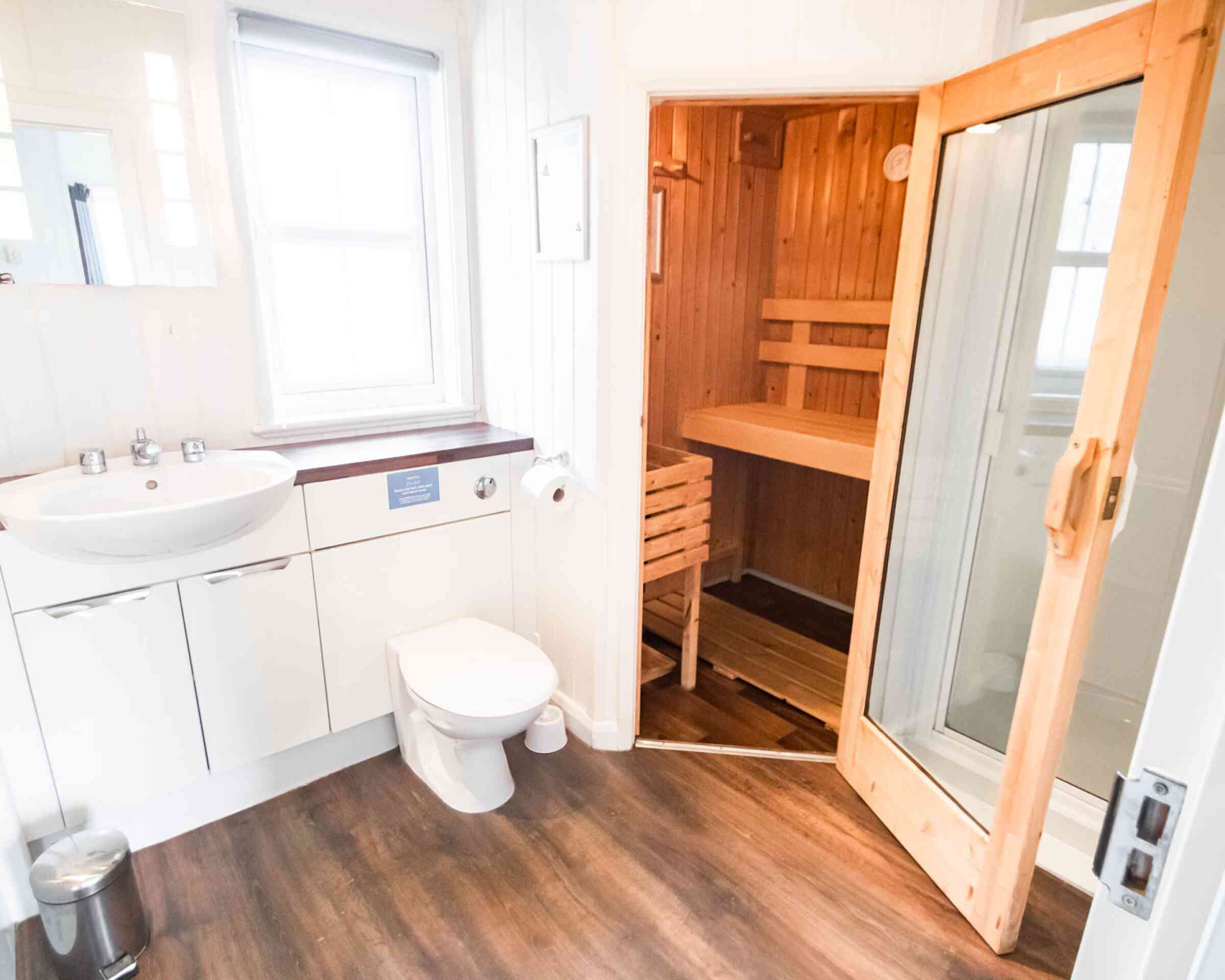 A modern bathroom featuring a white sink, toilet, and a wooden sauna visible through an open door. The room has light walls and a wooden floor, with natural light coming from a window.
