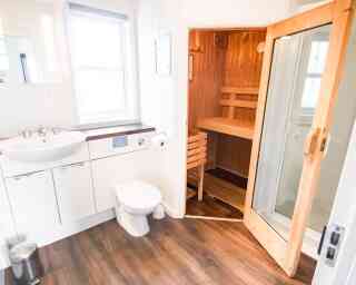 A modern bathroom featuring a white sink, toilet, and a wooden sauna visible through an open door. The room has light walls and a wooden floor, with natural light coming from a window.