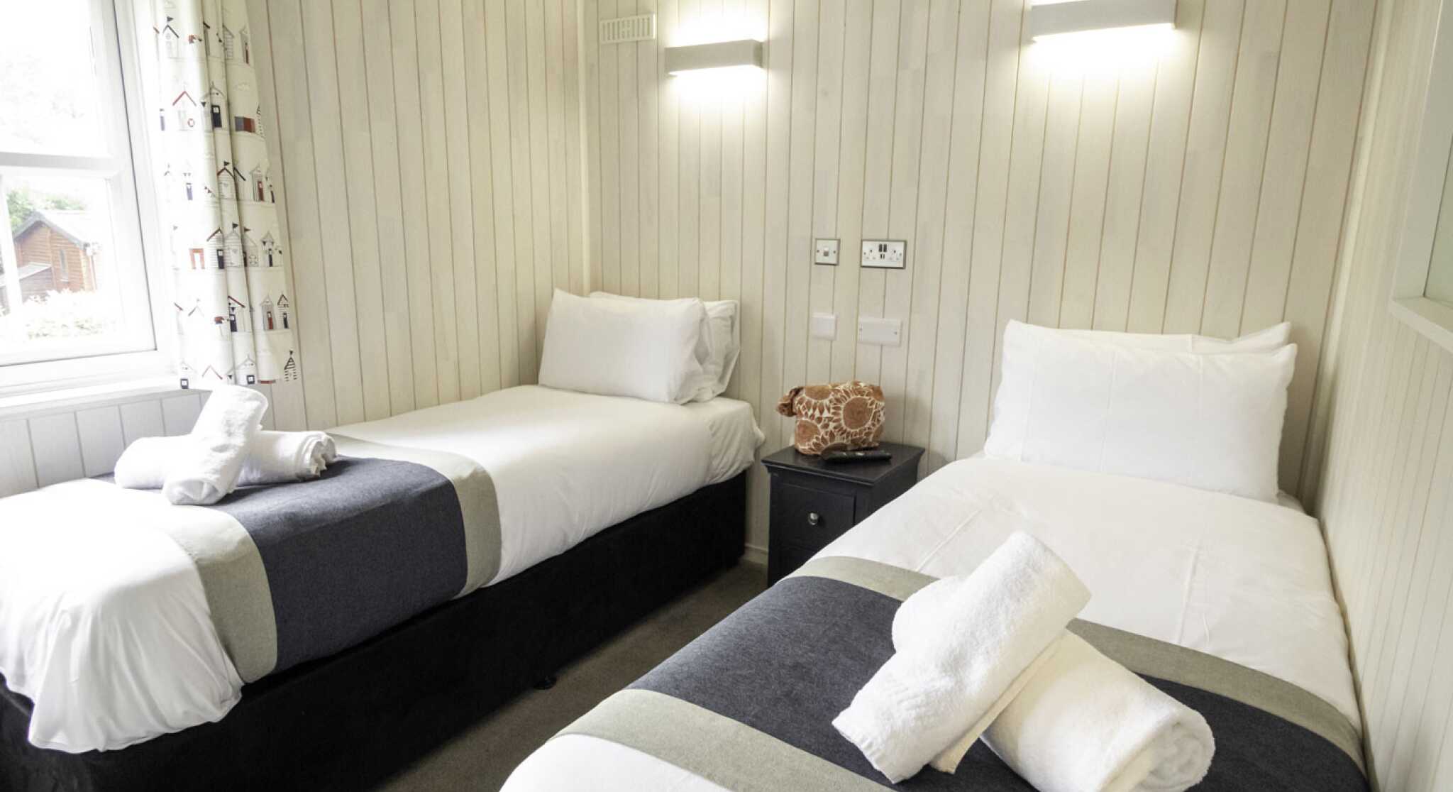 A cozy bedroom featuring two single beds with white linens and gray accents. Each bed is neatly made with rolled towels placed on top. The walls are lined with pale wood paneling, and there’s a bright window with light filtering through. A small nightstand sits between the beds, with a decorative coral piece on top.