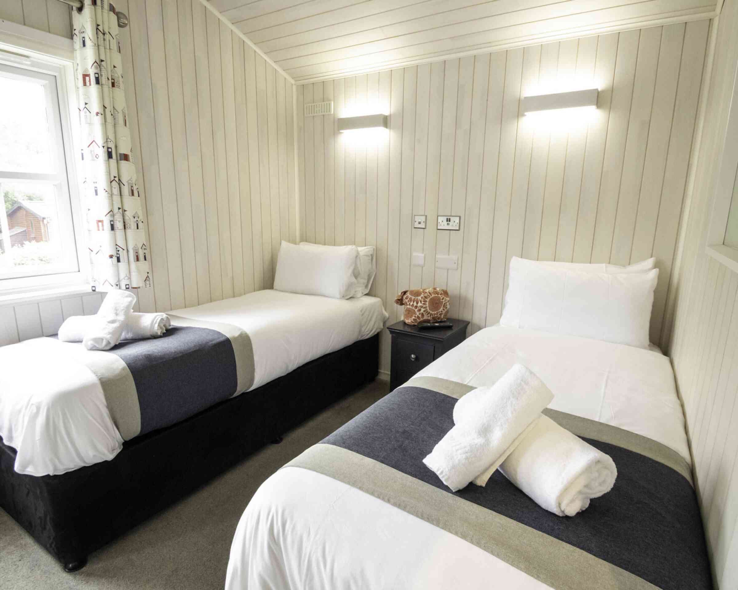 A cozy bedroom featuring two single beds with white linens and gray accents. Each bed is neatly made with rolled towels placed on top. The walls are lined with pale wood paneling, and there’s a bright window with light filtering through. A small nightstand sits between the beds, with a decorative coral piece on top.