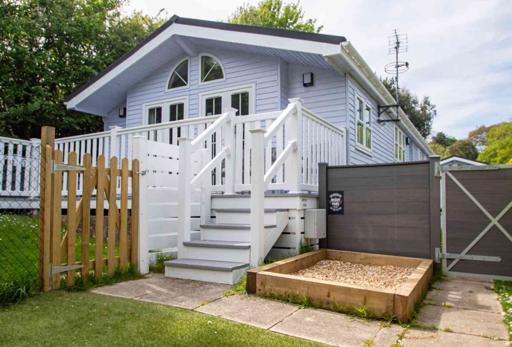 A light blue house with a white porch and balustrade, featuring a staircase leading to the entrance. There is a wooden fence and gate in the foreground, along with a small gravel area beside the house. Lush greenery surrounds the property.
