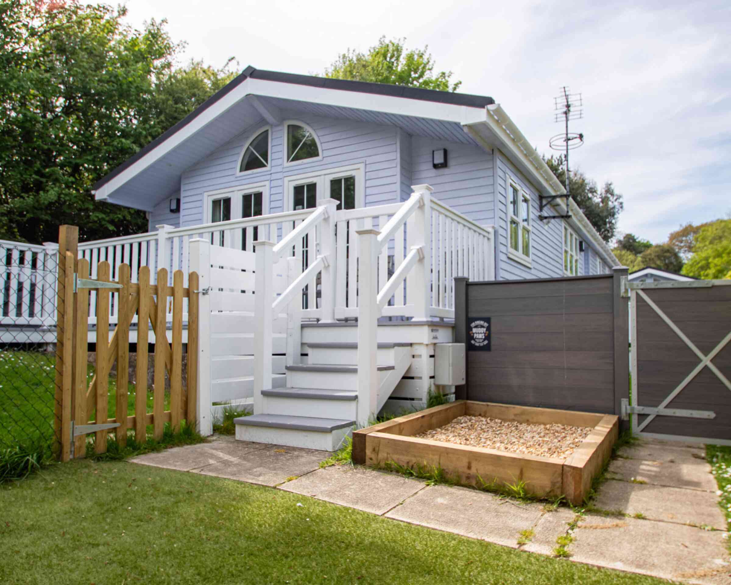 A light blue house with a white porch and balustrade, featuring a staircase leading to the entrance. There is a wooden fence and gate in the foreground, along with a small gravel area beside the house. Lush greenery surrounds the property.