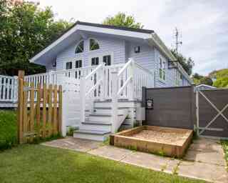 A light blue house with a white porch and balustrade, featuring a staircase leading to the entrance. There is a wooden fence and gate in the foreground, along with a small gravel area beside the house. Lush greenery surrounds the property.