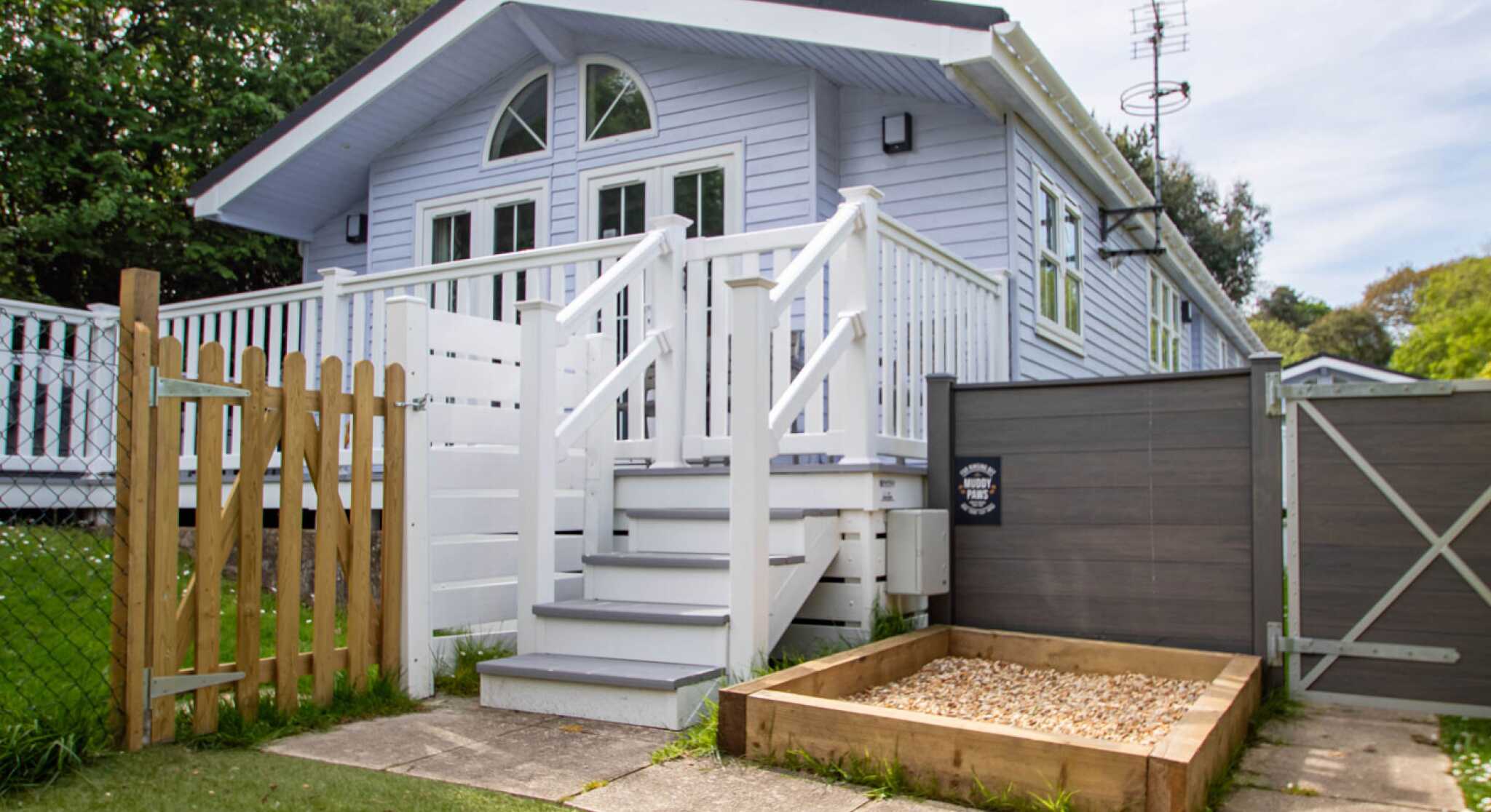 A light blue house with a white porch and balustrade, featuring a staircase leading to the entrance. There is a wooden fence and gate in the foreground, along with a small gravel area beside the house. Lush greenery surrounds the property.