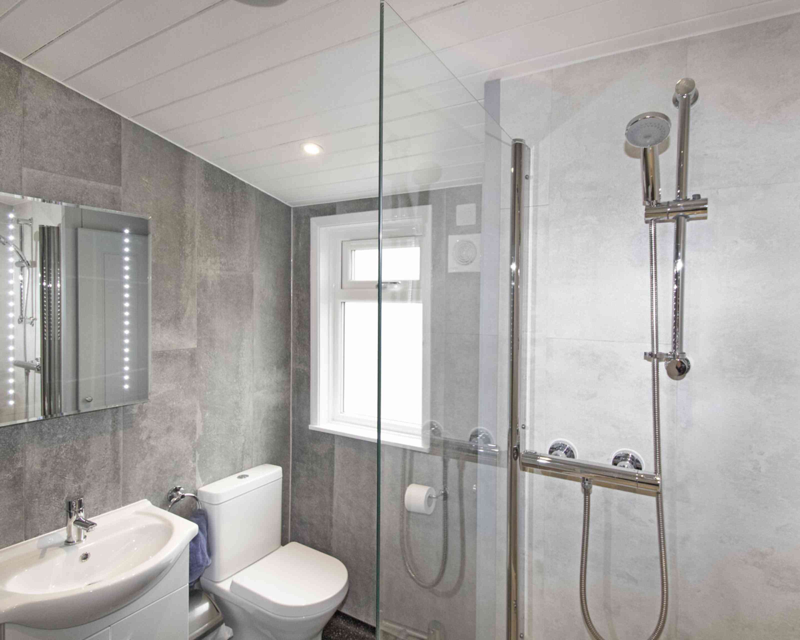 Modern bathroom featuring a glass shower enclosure, a sleek toilet, and a washbasin with a large mirror. The walls are tiled in grey, and there is natural light coming through a window.