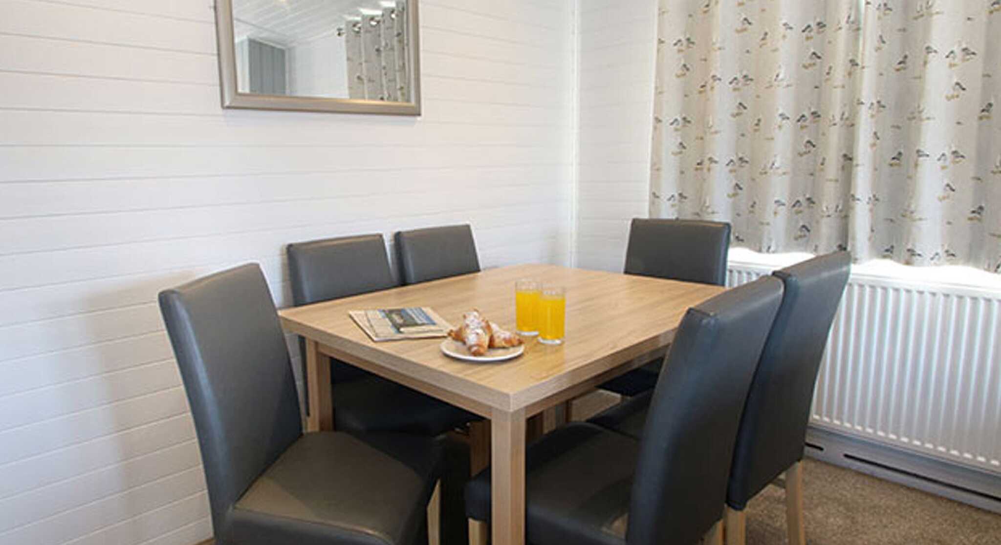 A modern dining area with a wooden table surrounded by six leather chairs. On the table, there are two glasses of orange juice and a plate with pastries. A mirror is mounted on the wall, and light curtains hang by the window.