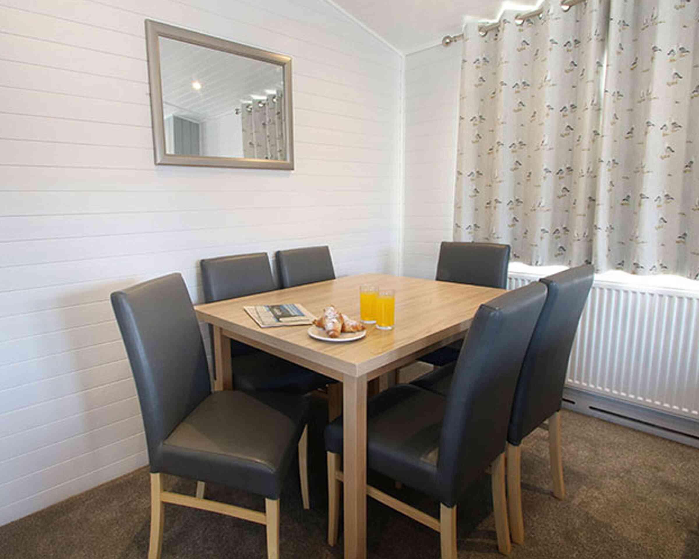 A modern dining area with a wooden table surrounded by six leather chairs. On the table, there are two glasses of orange juice and a plate with pastries. A mirror is mounted on the wall, and light curtains hang by the window.