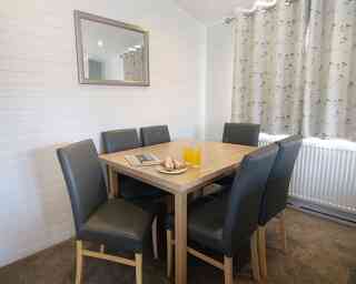 A modern dining area with a wooden table surrounded by six leather chairs. On the table, there are two glasses of orange juice and a plate with pastries. A mirror is mounted on the wall, and light curtains hang by the window.