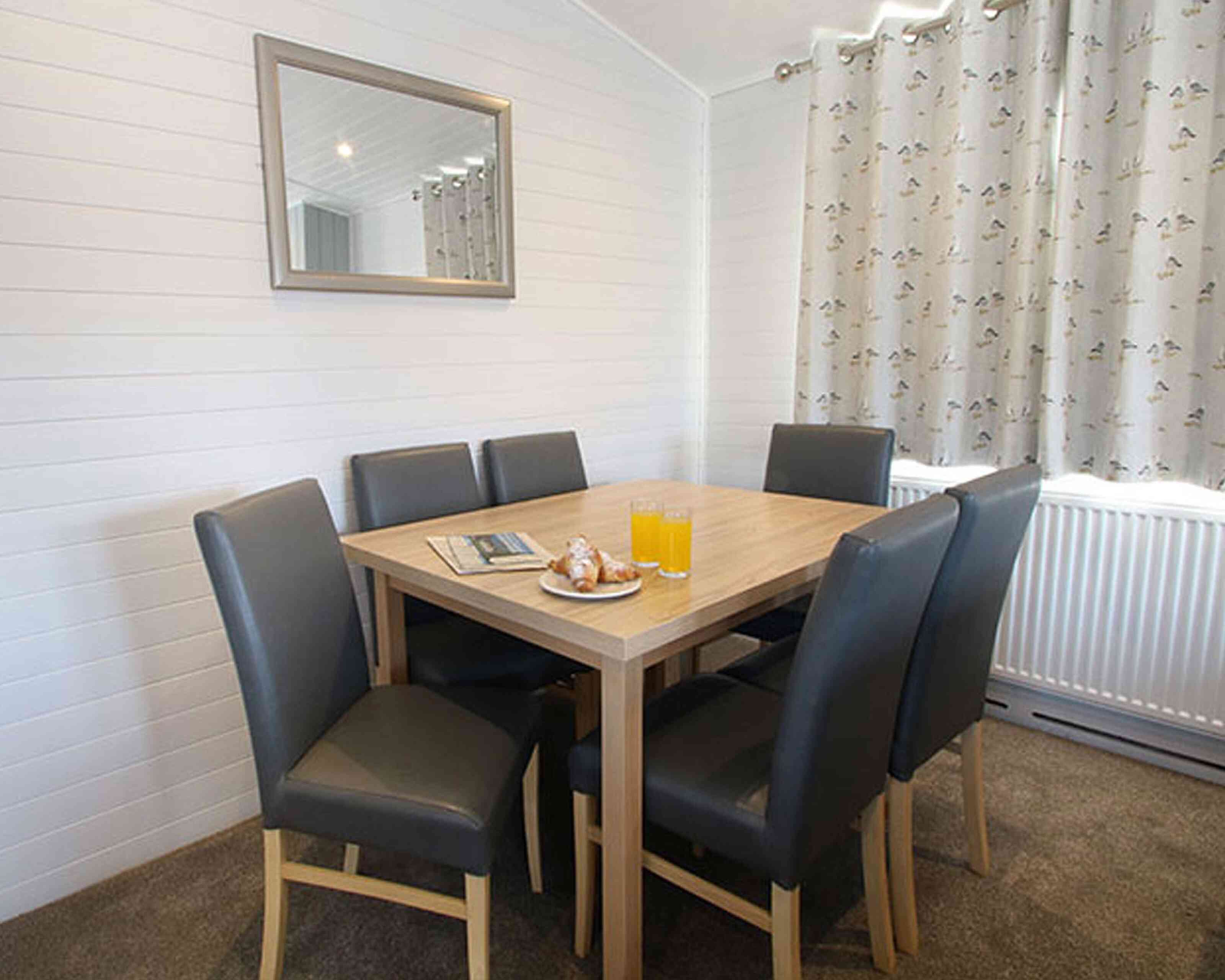 A modern dining area with a wooden table surrounded by six leather chairs. On the table, there are two glasses of orange juice and a plate with pastries. A mirror is mounted on the wall, and light curtains hang by the window.