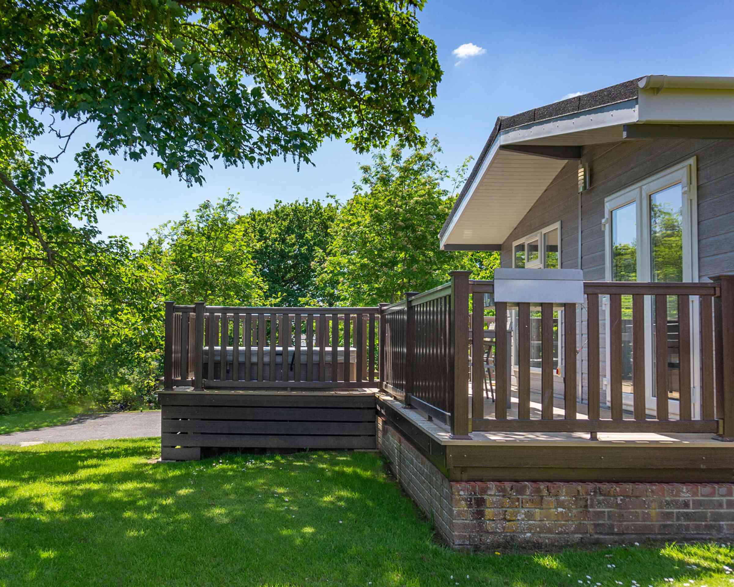 A wooden deck attached to a modern house, surrounded by lush green trees and grass under a clear blue sky. The deck features a railing and steps leading down to the yard.