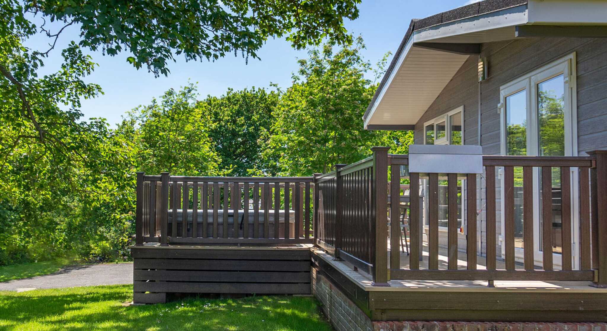 A wooden deck attached to a modern house, surrounded by lush green trees and grass under a clear blue sky. The deck features a railing and steps leading down to the yard.