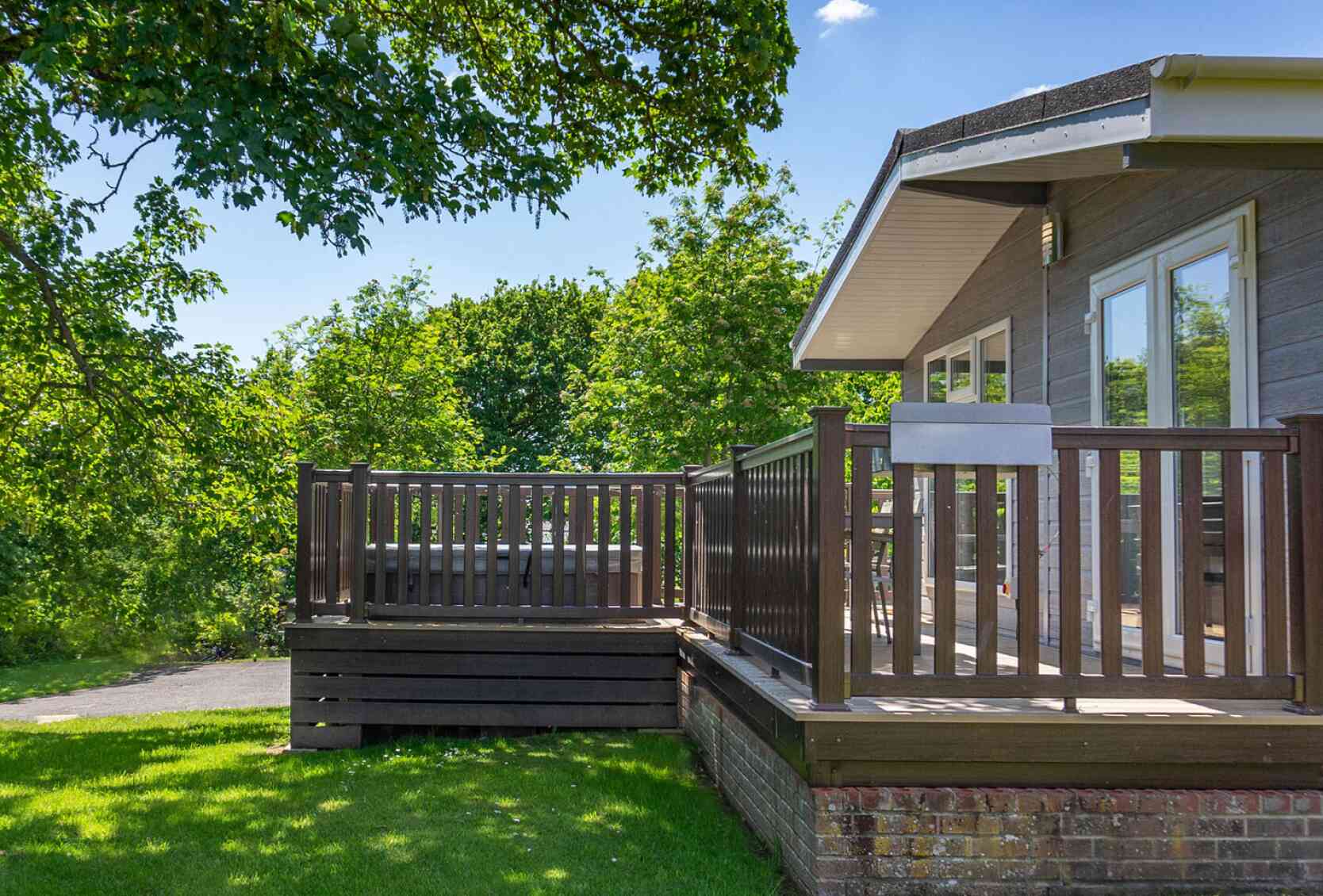 A wooden deck attached to a modern house, surrounded by lush green trees and grass under a clear blue sky. The deck features a railing and steps leading down to the yard.