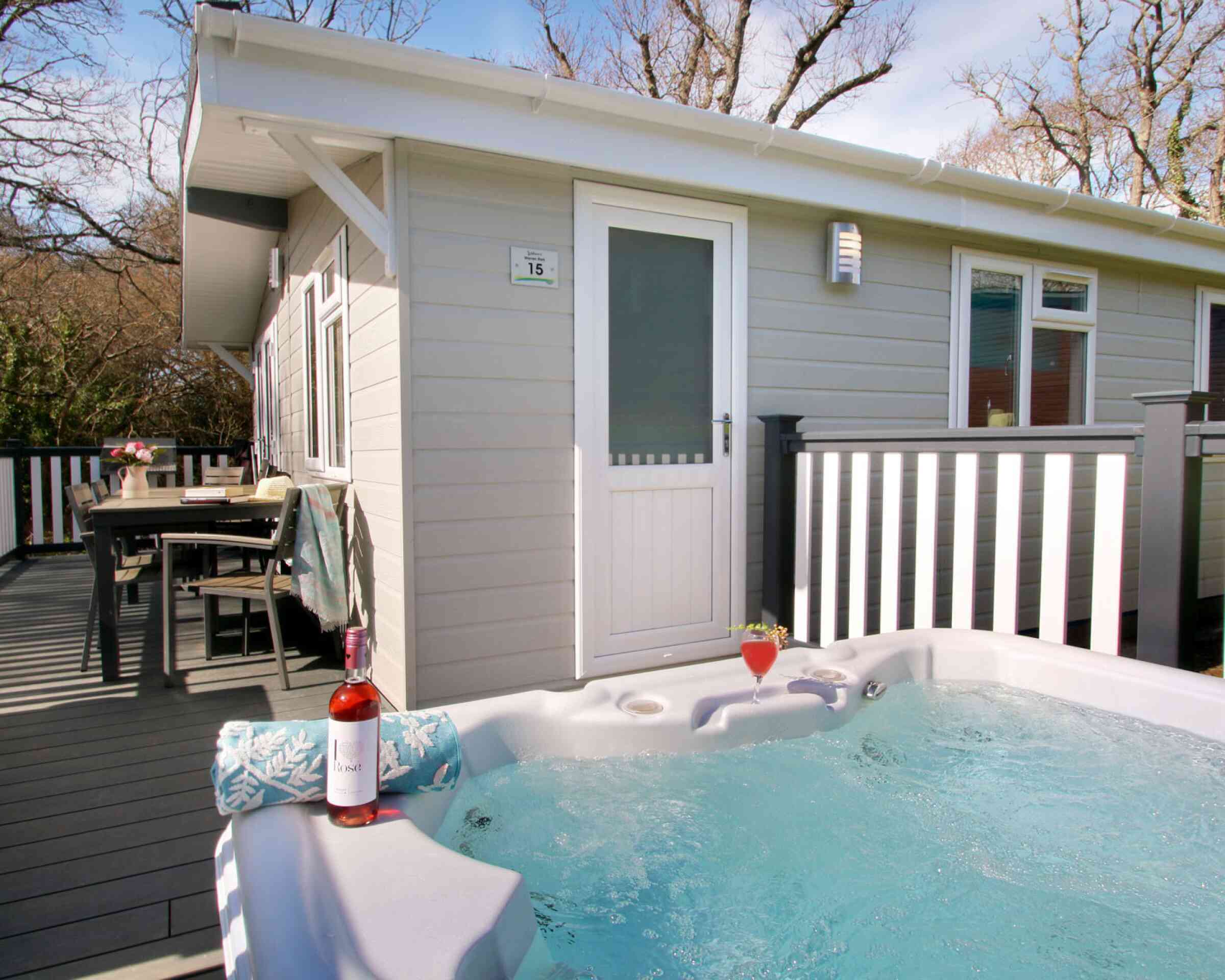 A hot tub filled with water sits on a wooden deck in front of a modern cabin. A bottle of rose wine and a glass are placed on the edge of the hot tub. Nearby, a dining table is set outside, surrounded by trees. The cabin features a white door and multiple windows.