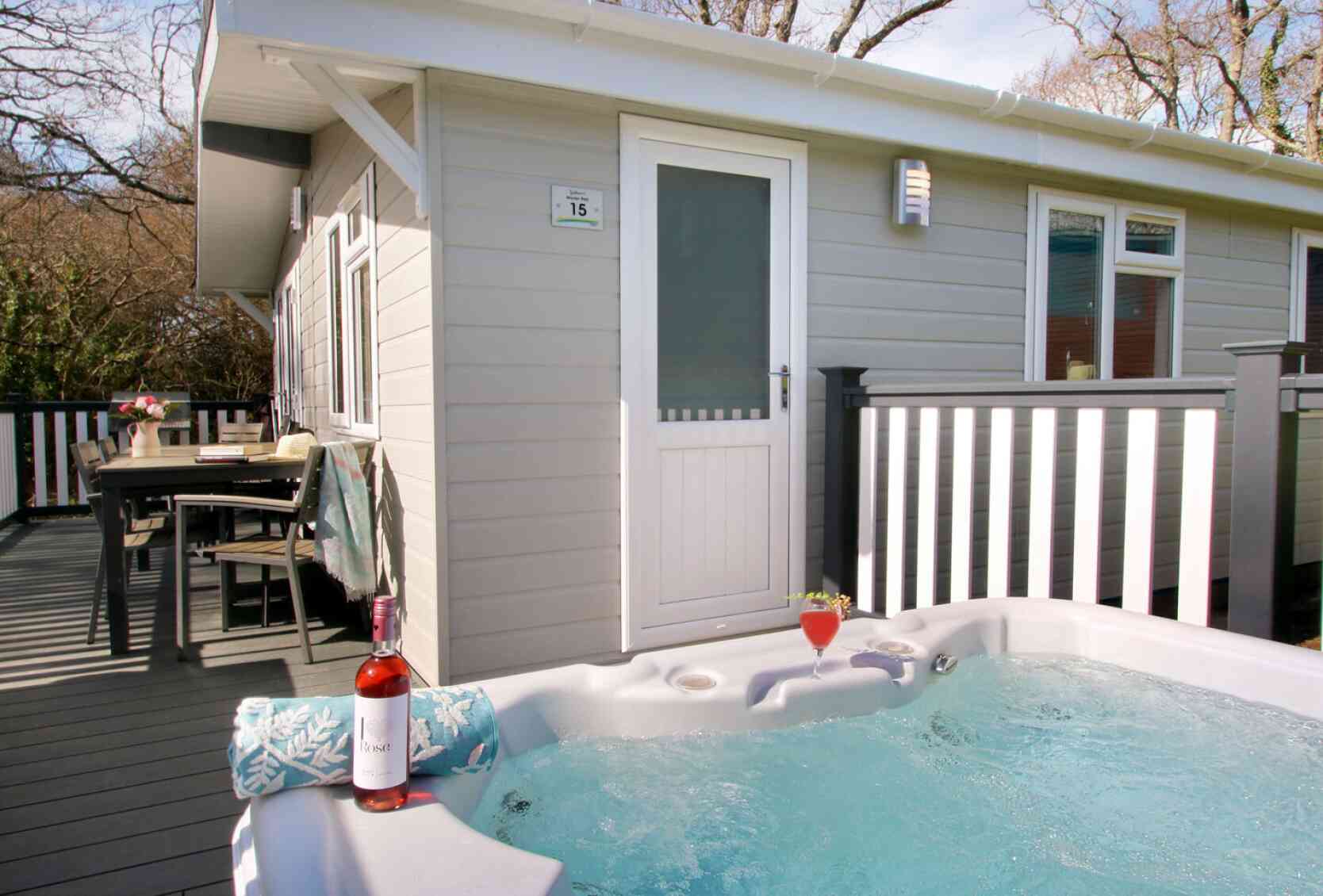 A hot tub filled with water sits on a wooden deck in front of a modern cabin. A bottle of rose wine and a glass are placed on the edge of the hot tub. Nearby, a dining table is set outside, surrounded by trees. The cabin features a white door and multiple windows.