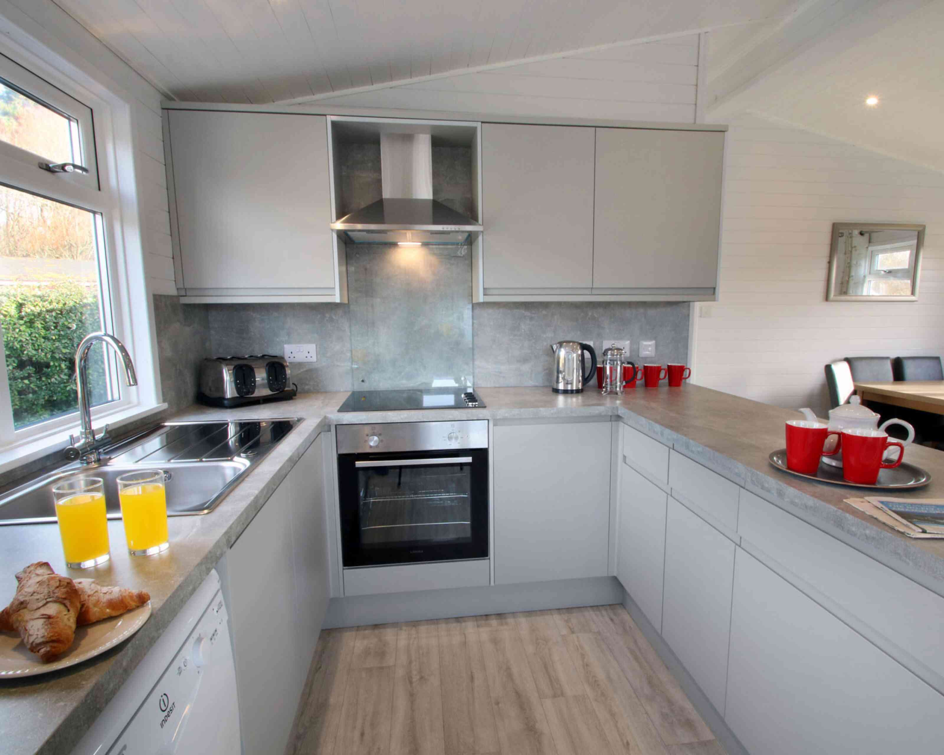 A modern kitchen featuring gray cabinets, a stainless steel oven, and a sleek countertop. Two glasses of orange juice are on the counter, alongside croissants and red mugs. Natural light illuminates the space through a window.