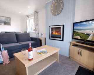 A cozy living room featuring a gray sofa, a wooden coffee table with a bottle of wine and two glasses, a magazine, and a television displaying a scenic view. A modern clock hangs on the wall above a sleek electric fireplace, creating a warm atmosphere.