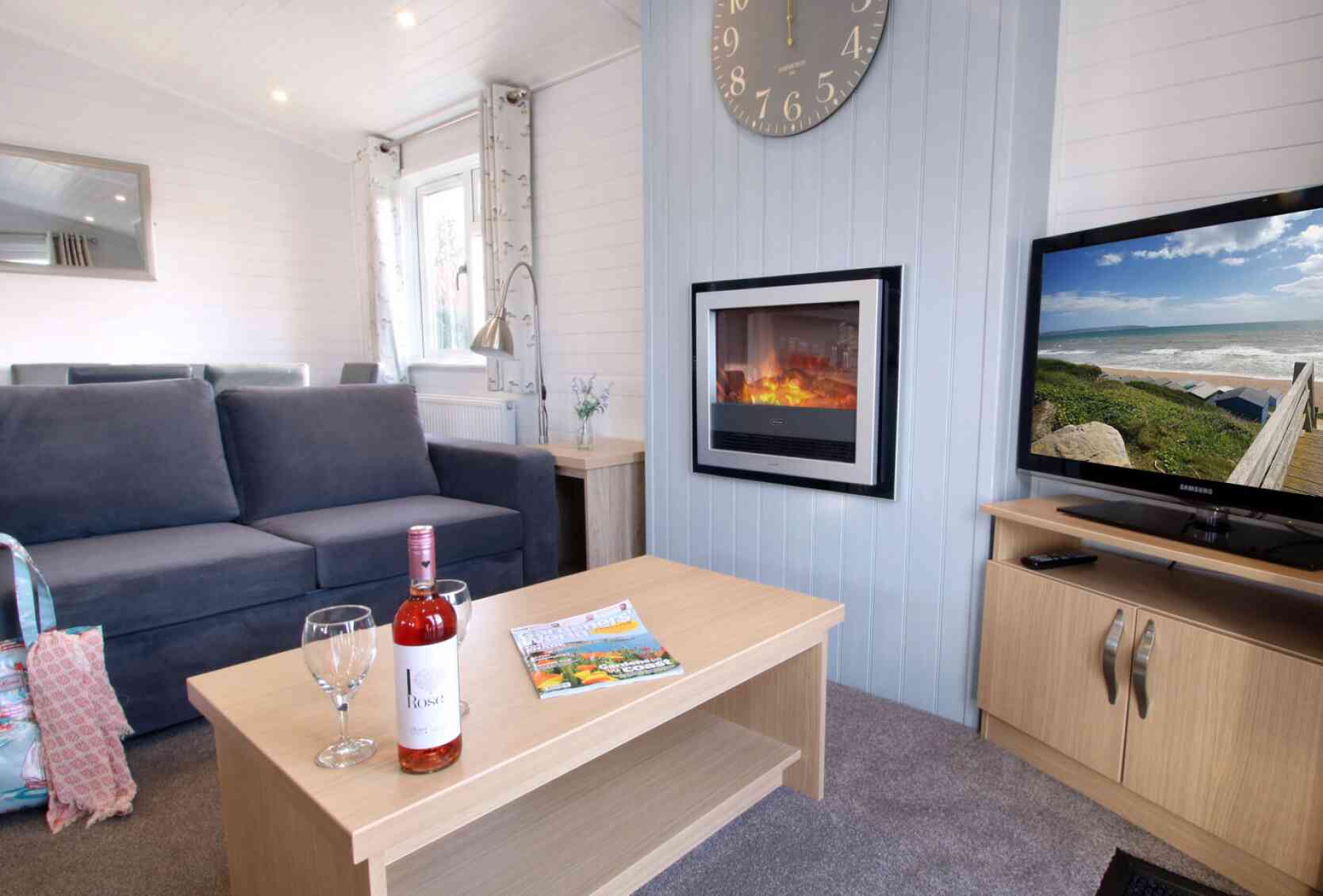 A cozy living room featuring a gray sofa, a wooden coffee table with a bottle of wine and two glasses, a magazine, and a television displaying a scenic view. A modern clock hangs on the wall above a sleek electric fireplace, creating a warm atmosphere.