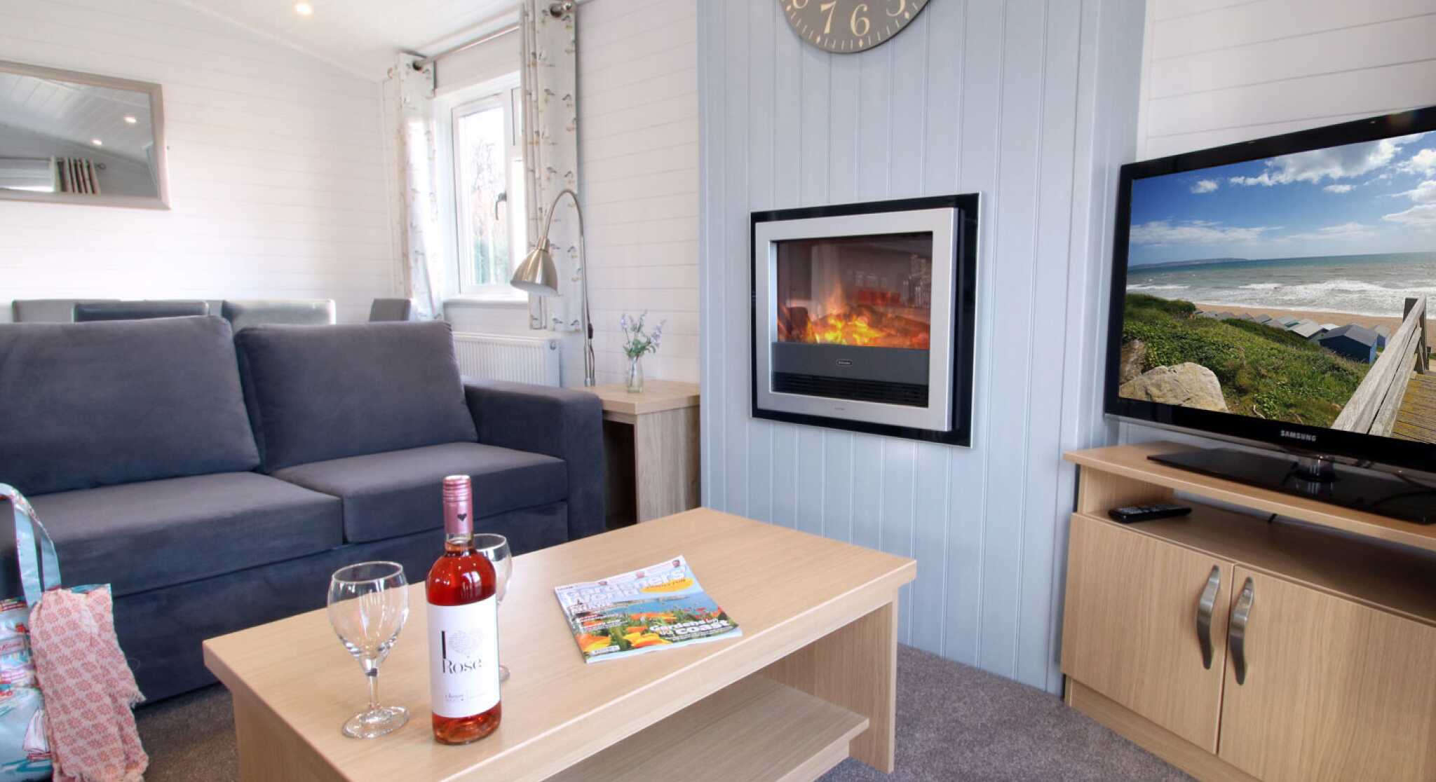 A cozy living room featuring a gray sofa, a wooden coffee table with a bottle of wine and two glasses, a magazine, and a television displaying a scenic view. A modern clock hangs on the wall above a sleek electric fireplace, creating a warm atmosphere.