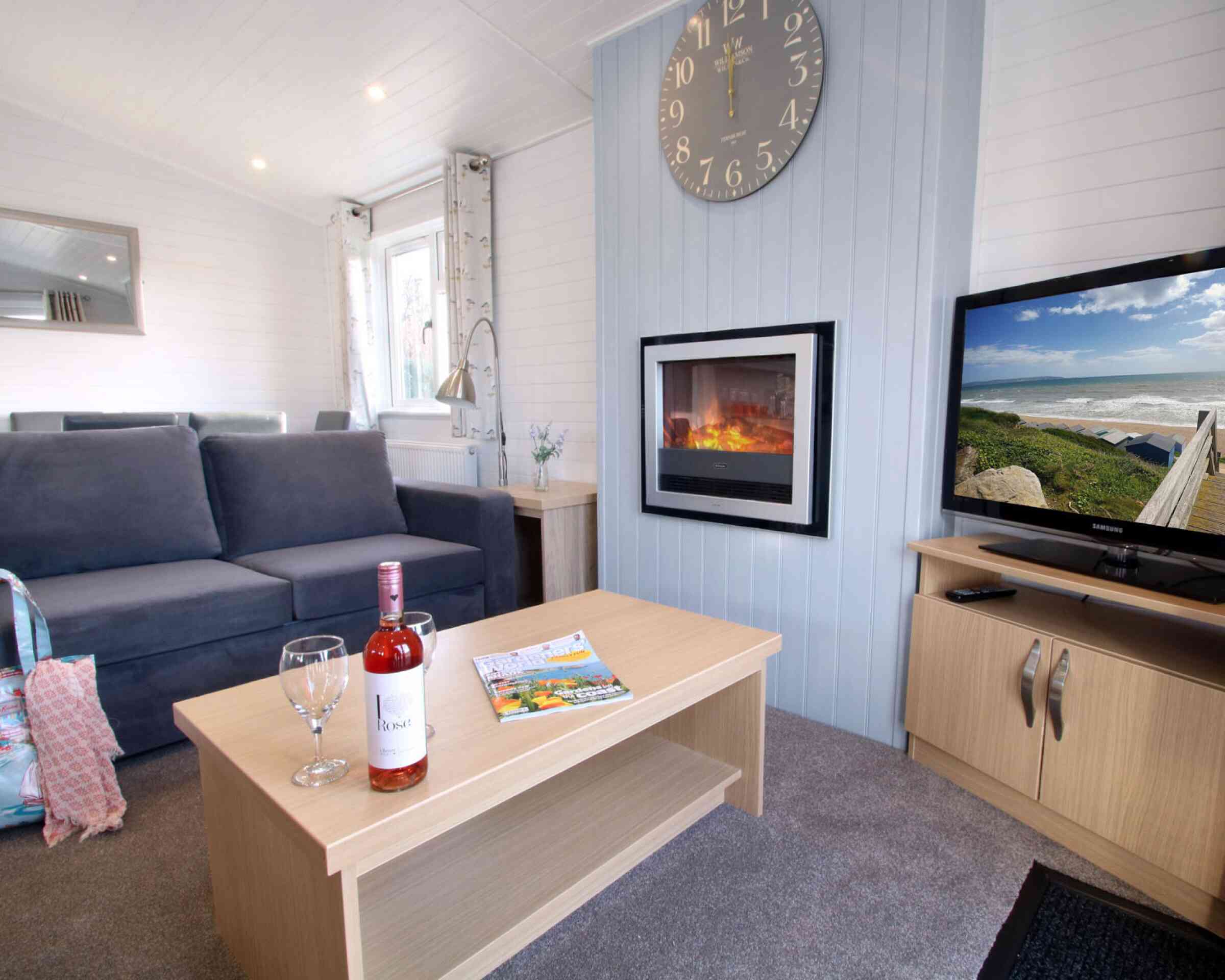 A cozy living room featuring a gray sofa, a wooden coffee table with a bottle of wine and two glasses, a magazine, and a television displaying a scenic view. A modern clock hangs on the wall above a sleek electric fireplace, creating a warm atmosphere.