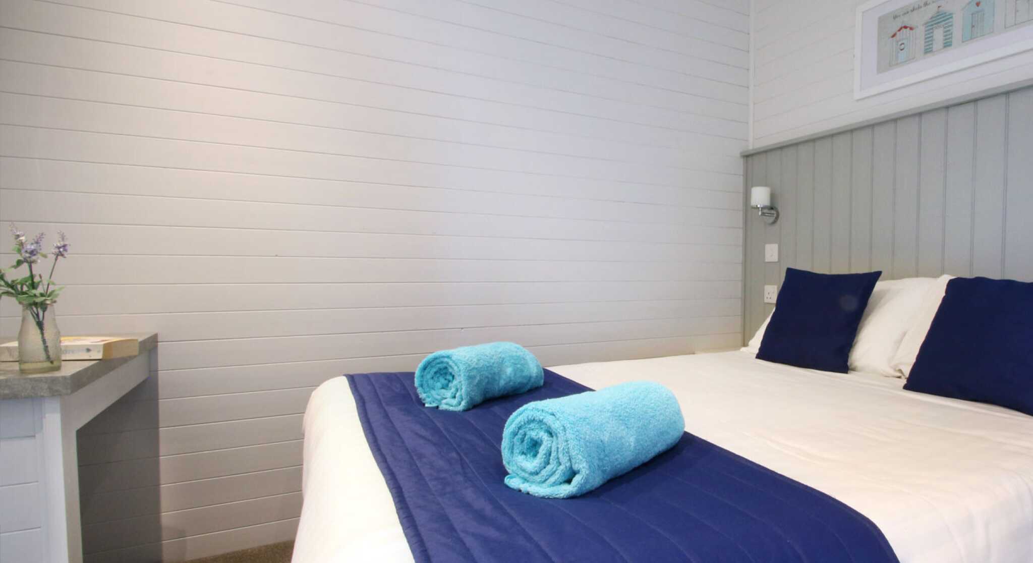 A cozy bedroom featuring a large bed with white linens and navy blue accents. Two turquoise towels are neatly rolled on the bed. A simple wooden table is in the corner, and the walls have a light gray wood paneling.