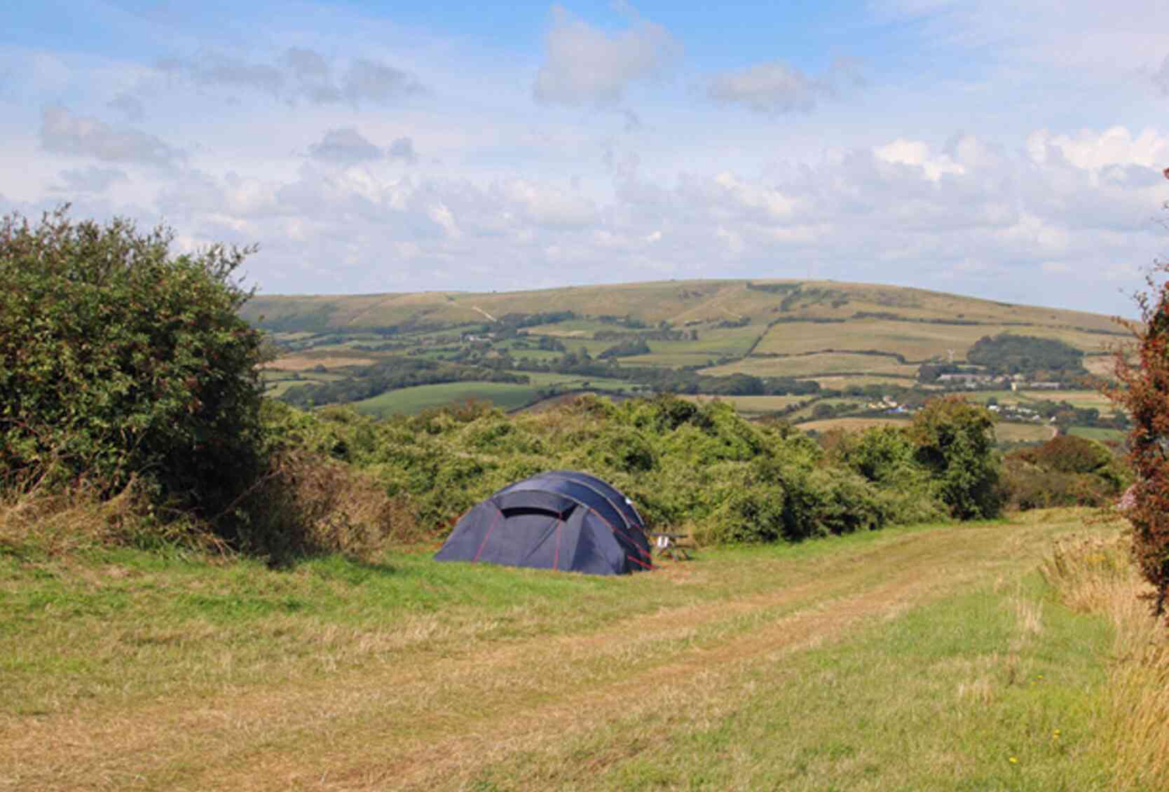 A blue tent is set up on a grassy hill with rolling green hills and a blue sky in the background. Fluffy white clouds drift across the sky, creating a serene countryside atmosphere.