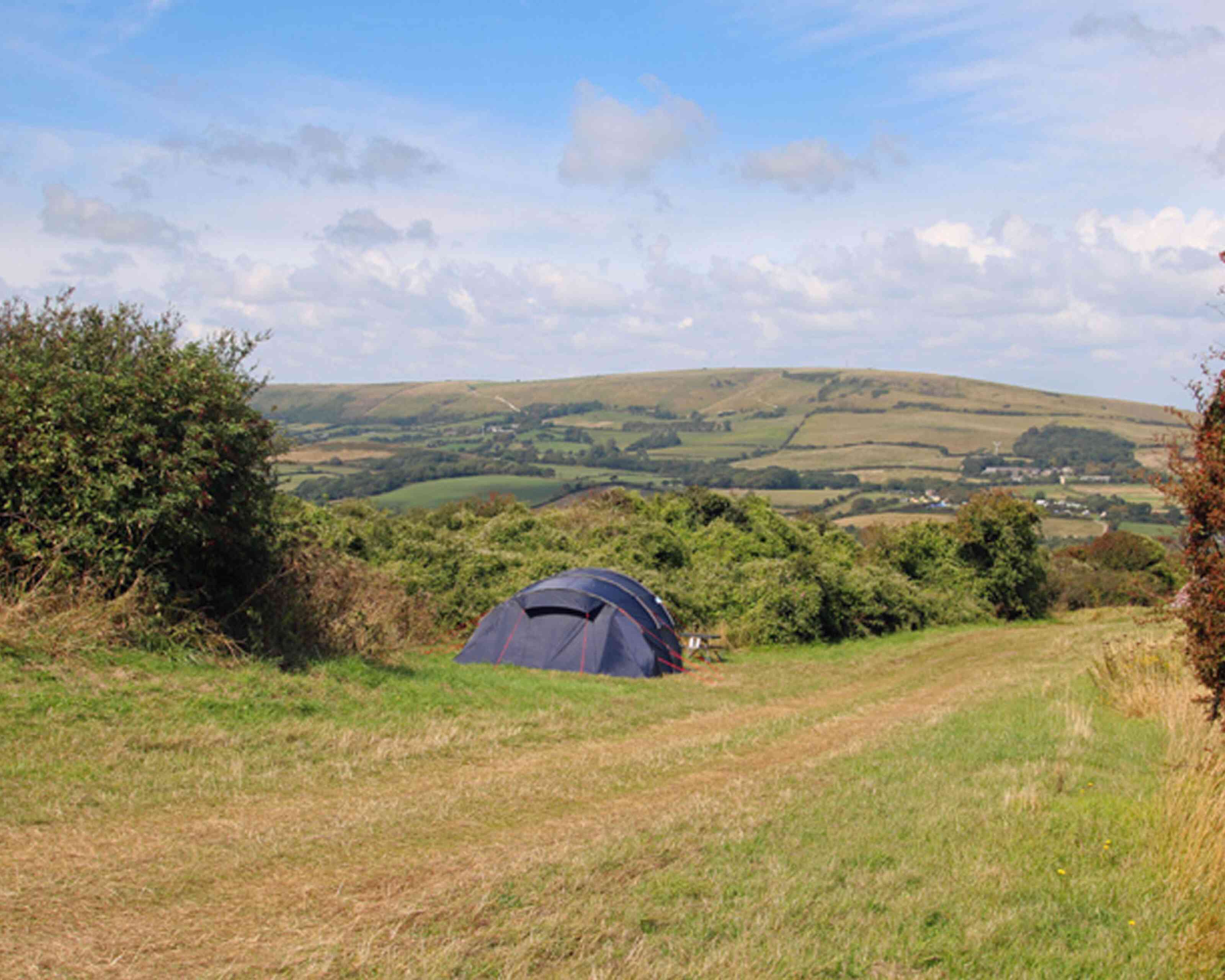 A blue tent is set up on a grassy hill with rolling green hills and a blue sky in the background. Fluffy white clouds drift across the sky, creating a serene countryside atmosphere.