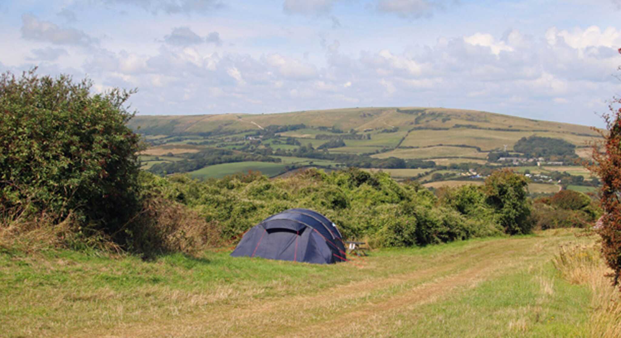 A blue tent is set up on a grassy hill with rolling green hills and a blue sky in the background. Fluffy white clouds drift across the sky, creating a serene countryside atmosphere.