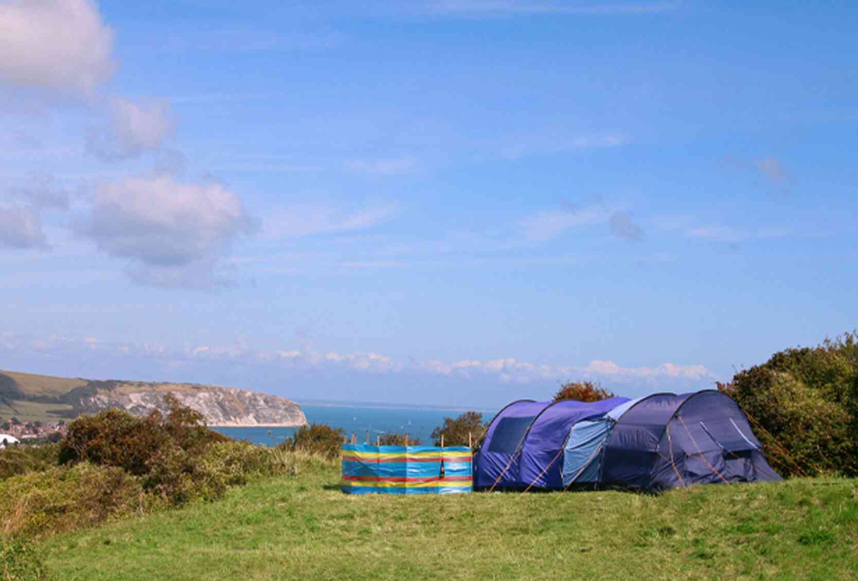 Camping tents set up on a grassy hill overlooking a scenic coastline. The ocean is visible in the background under a clear blue sky.