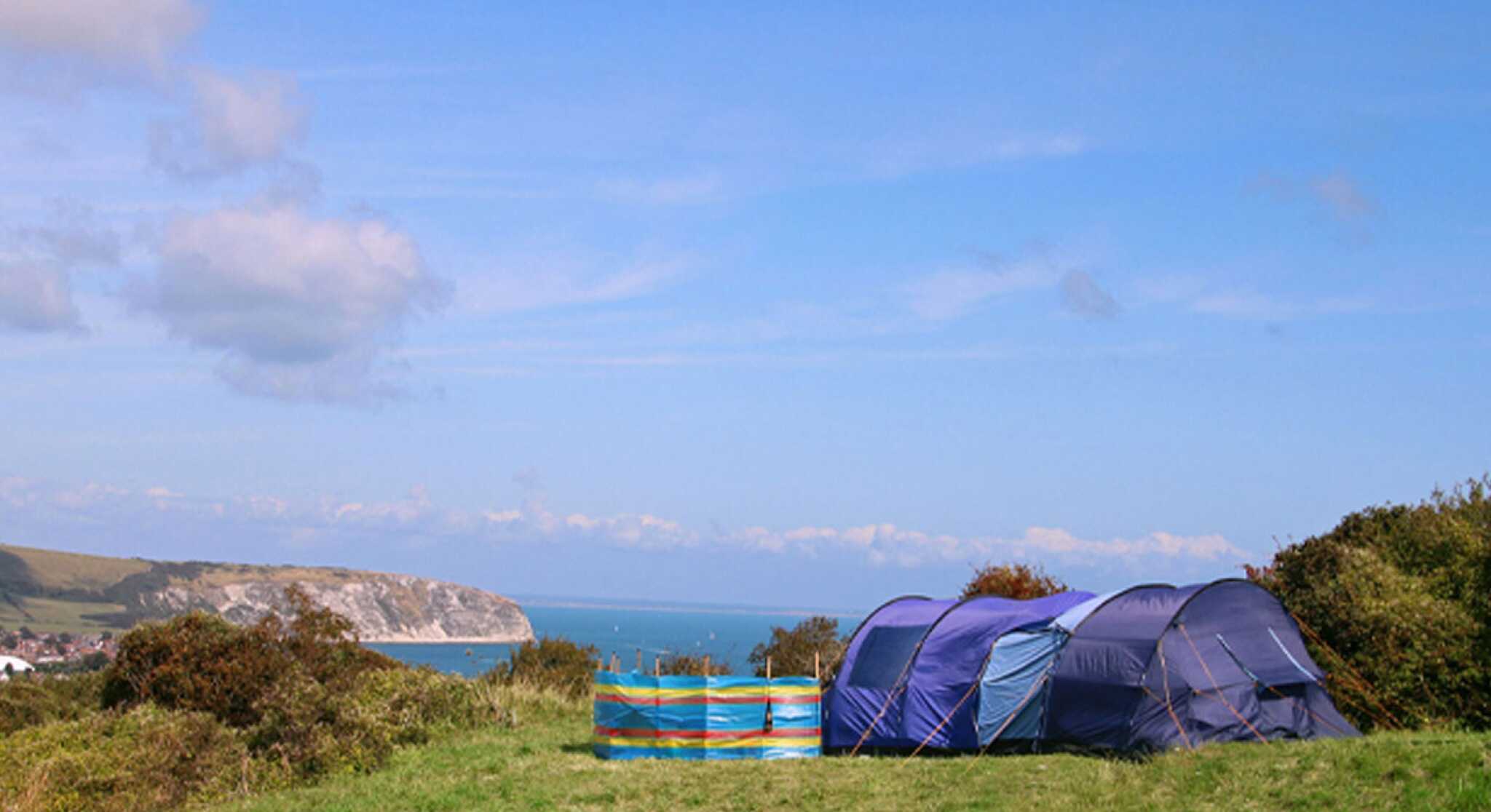 Camping tents set up on a grassy hill overlooking a scenic coastline. The ocean is visible in the background under a clear blue sky.
