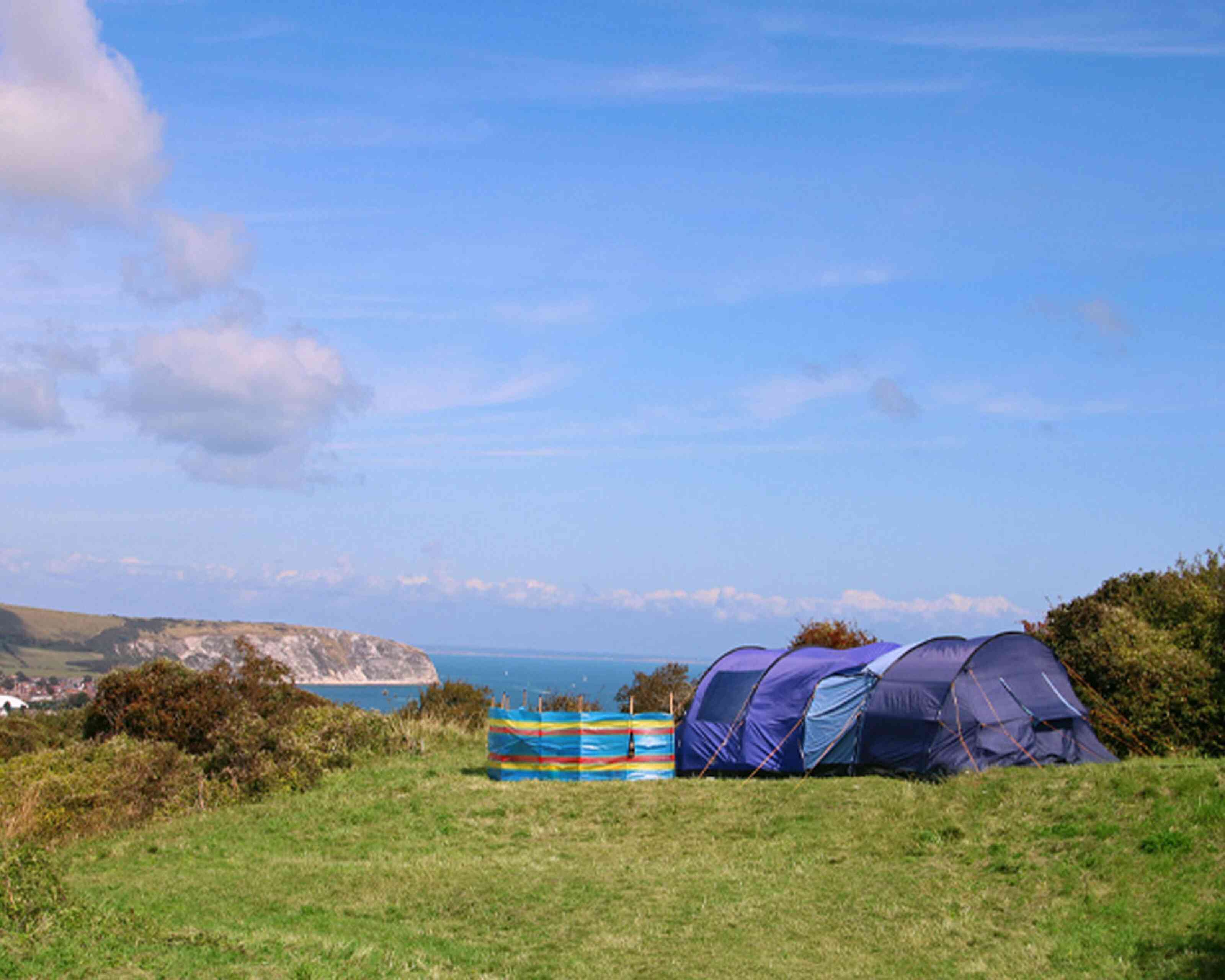 Camping tents set up on a grassy hill overlooking a scenic coastline. The ocean is visible in the background under a clear blue sky.