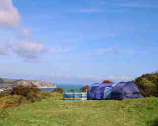 Camping tents set up on a grassy hill overlooking a scenic coastline. The ocean is visible in the background under a clear blue sky.