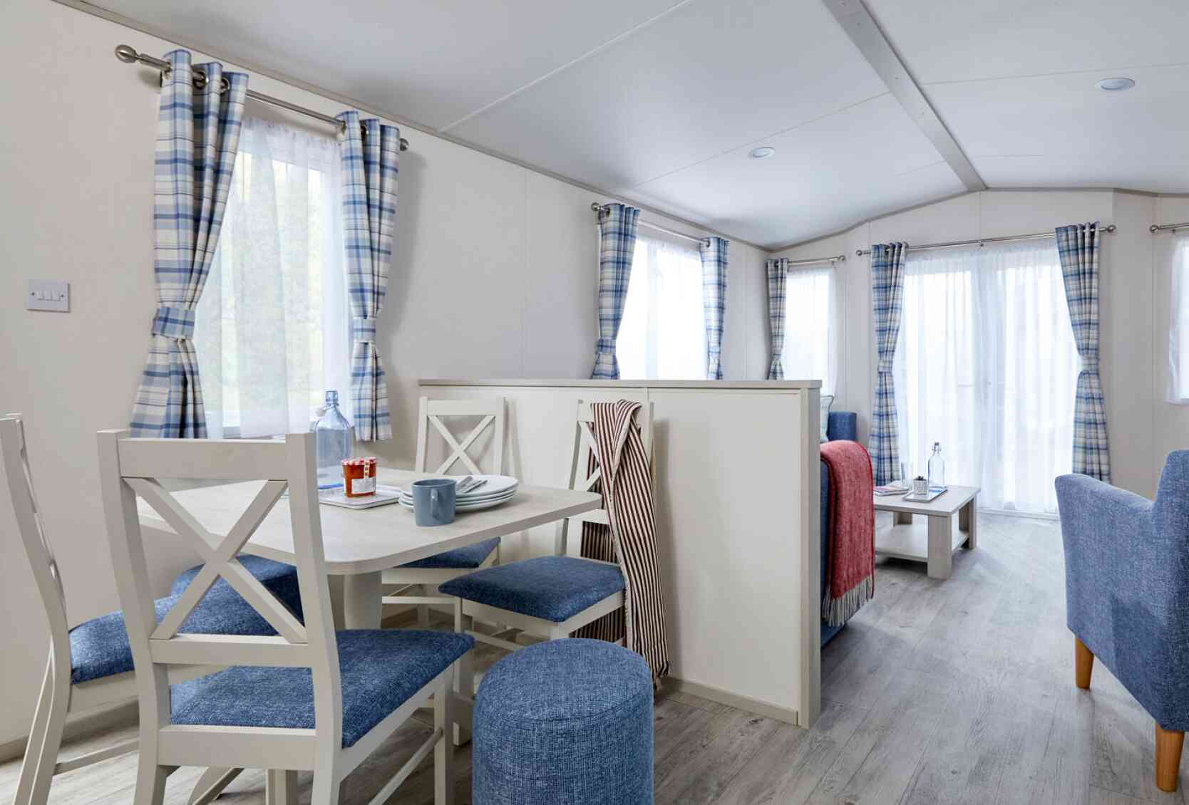 A cozy interior of a modern mobile home featuring a dining area with a round table and four blue chairs, a living space with a gray sofa and table, and large windows dressed with checked curtains. The wooden flooring enhances the bright and airy feel of the space.
