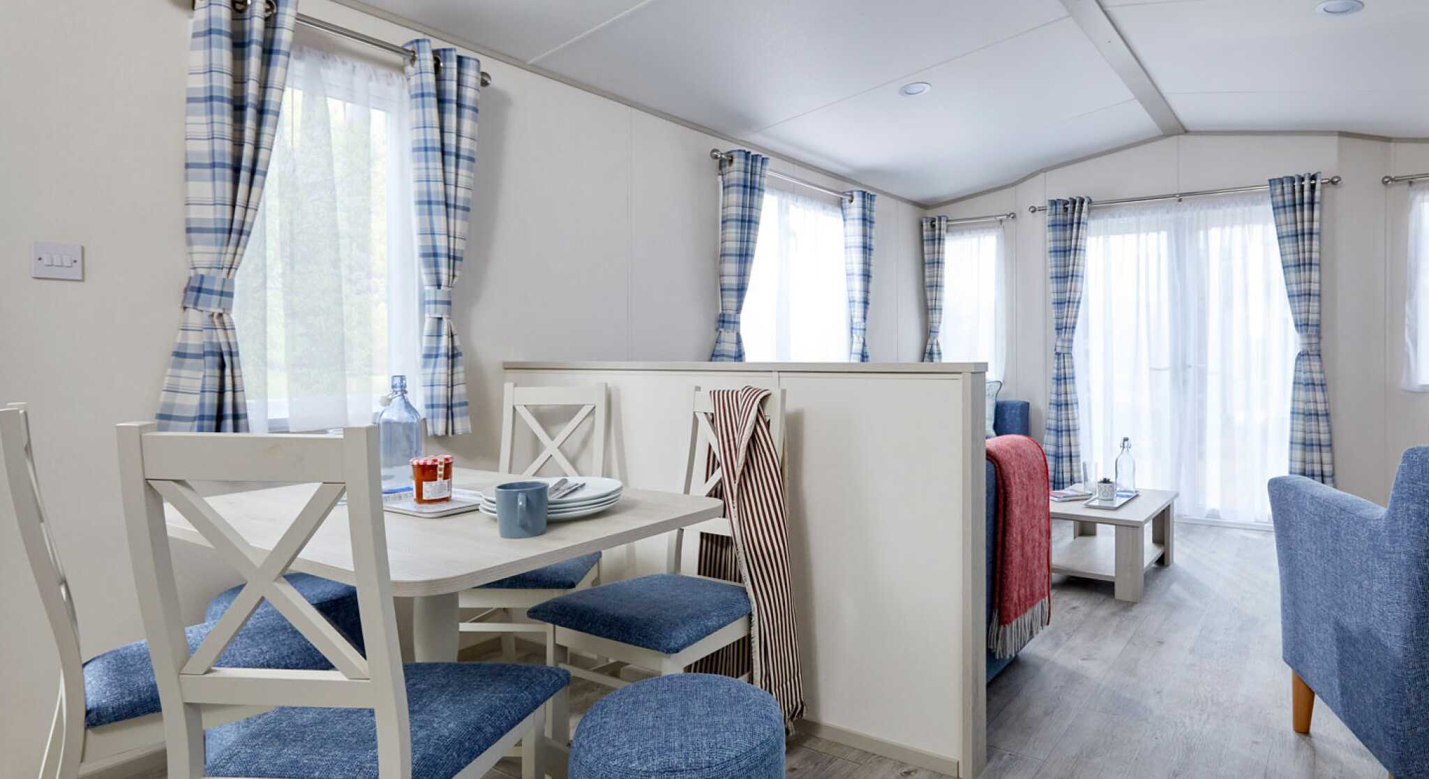 A cozy interior of a modern mobile home featuring a dining area with a round table and four blue chairs, a living space with a gray sofa and table, and large windows dressed with checked curtains. The wooden flooring enhances the bright and airy feel of the space.