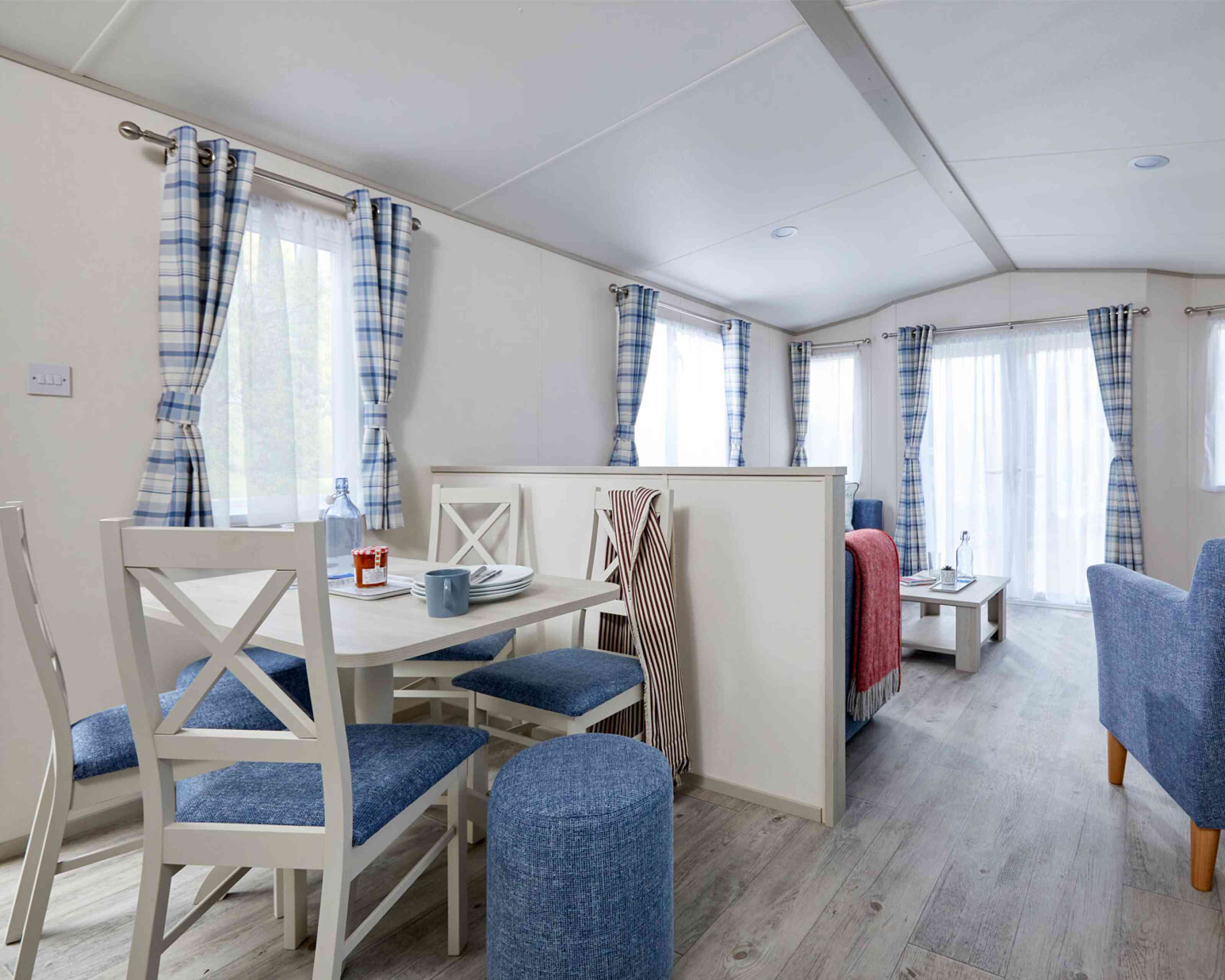 A cozy interior of a modern mobile home featuring a dining area with a round table and four blue chairs, a living space with a gray sofa and table, and large windows dressed with checked curtains. The wooden flooring enhances the bright and airy feel of the space.