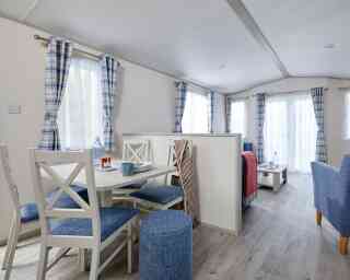 A cozy interior of a modern mobile home featuring a dining area with a round table and four blue chairs, a living space with a gray sofa and table, and large windows dressed with checked curtains. The wooden flooring enhances the bright and airy feel of the space.