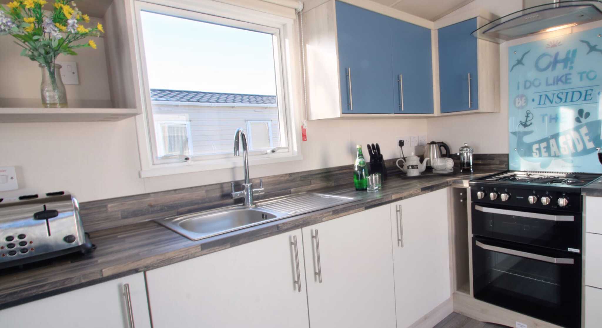A modern kitchen with light-colored cabinetry, blue accents, and a stainless steel sink. The kitchen includes a gas stove, coffee maker, toaster, and bottles on the counter. A window allows natural light to enter, enhancing the airy feel of the space. Decorative flowers are placed in a vase.