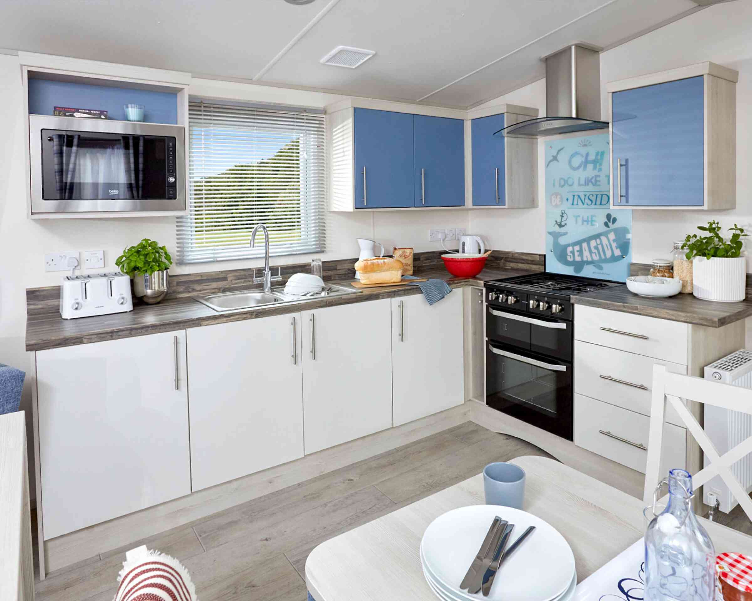 Modern kitchen with blue and white cabinetry, featuring a microwave, sink, and gas stove. A small dining table with plates and cups is visible in the foreground. Natural light streams through a window with sheer curtains.