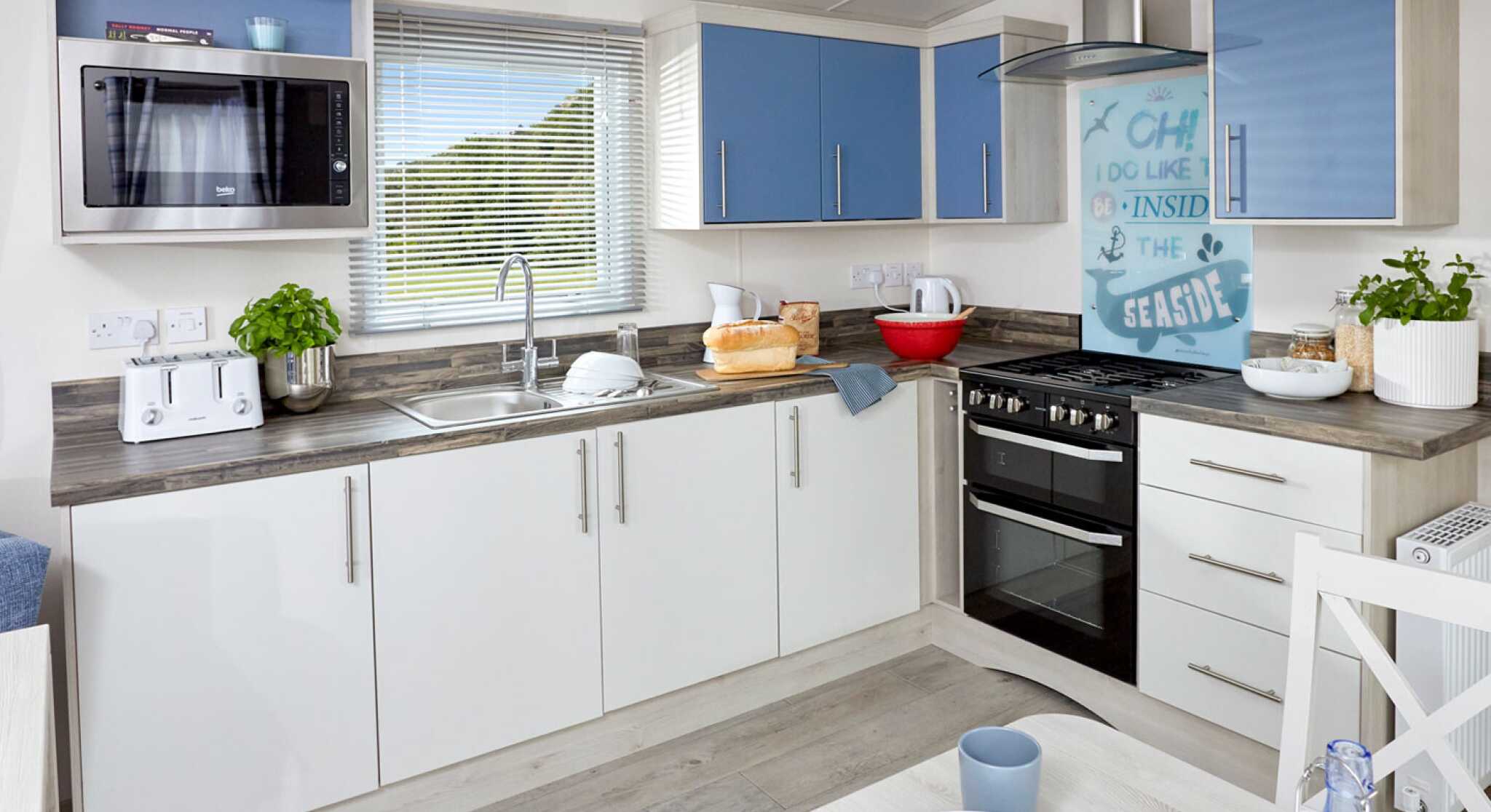 Modern kitchen with blue and white cabinetry, featuring a microwave, sink, and gas stove. A small dining table with plates and cups is visible in the foreground. Natural light streams through a window with sheer curtains.